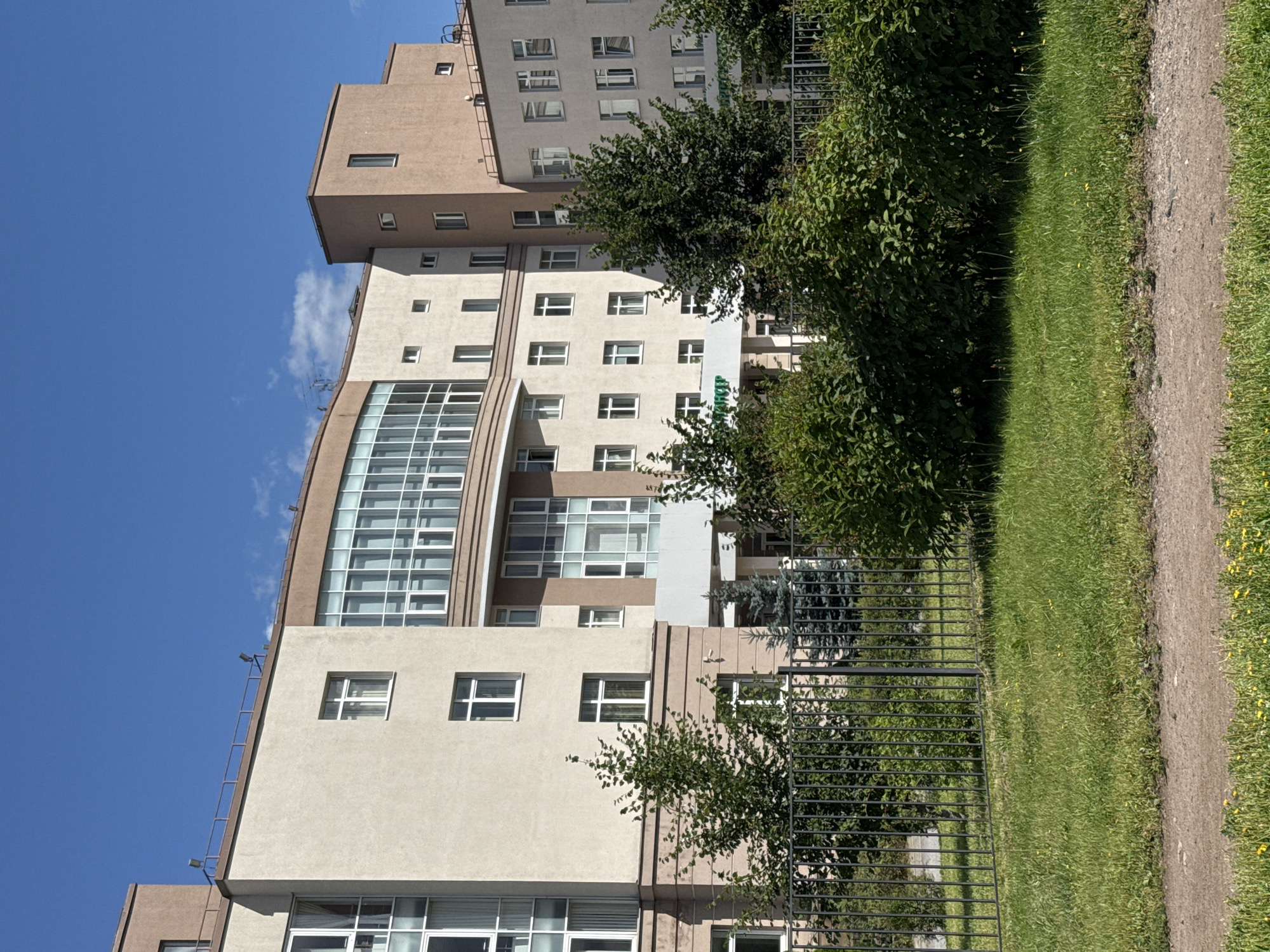 A sunlit, multi-story building with a modern architectural design stands under a clear blue sky. The building features a mix of beige and brown facade colors, with numerous windows, some of which are large and curved glass panels on the upper floors. A green sign with white lettering is visible above a covered entrance. In front of the building, there is a dark metal fence, behind which various trees and bushes are growing. The foreground shows a well-maintained lawn with a dirt path. The scene is consistent with the facade of the St. Petersburg State Transport University's dormitory building, located at 104 Bolshevikov Avenue, Saint Petersburg, Russia. There are no people or vehicles visible in the image.