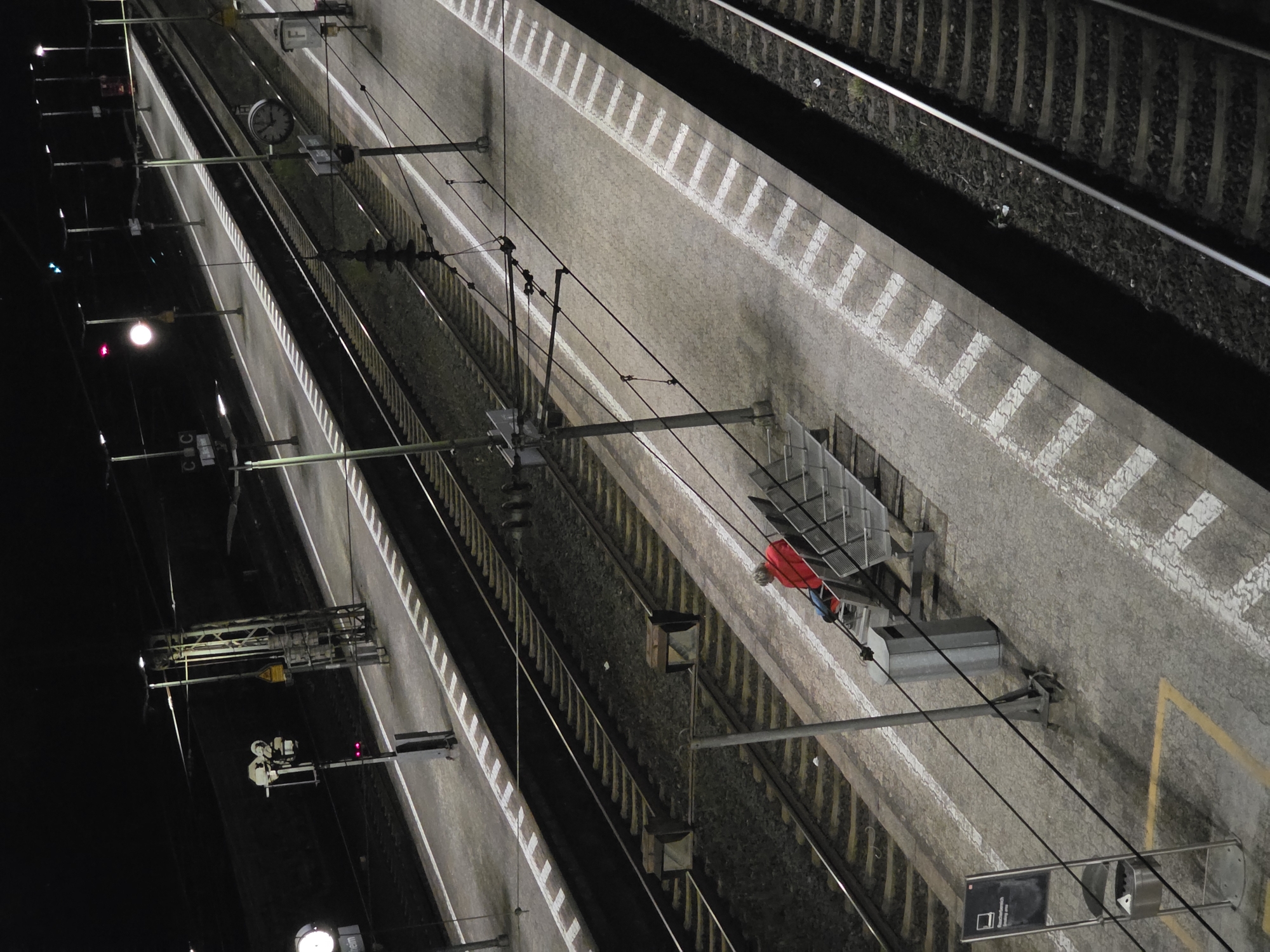 The image depicts a nighttime scene at the Bischofsheim train station in Germany. A single individual, wearing a red jacket and blue pants, is seated on a bench on a train platform. The platform is paved with textured concrete and marked with white lines. Multiple sets of train tracks run alongside the platform, illuminated by artificial lighting. Overhead power lines and signal lights are visible, typical of a train station environment. The overall atmosphere is quiet and somewhat desolate, with the individual appearing to be waiting. A trash receptacle and a sign labeled "Raucherbereich" (smoking area) are also present on the platform.