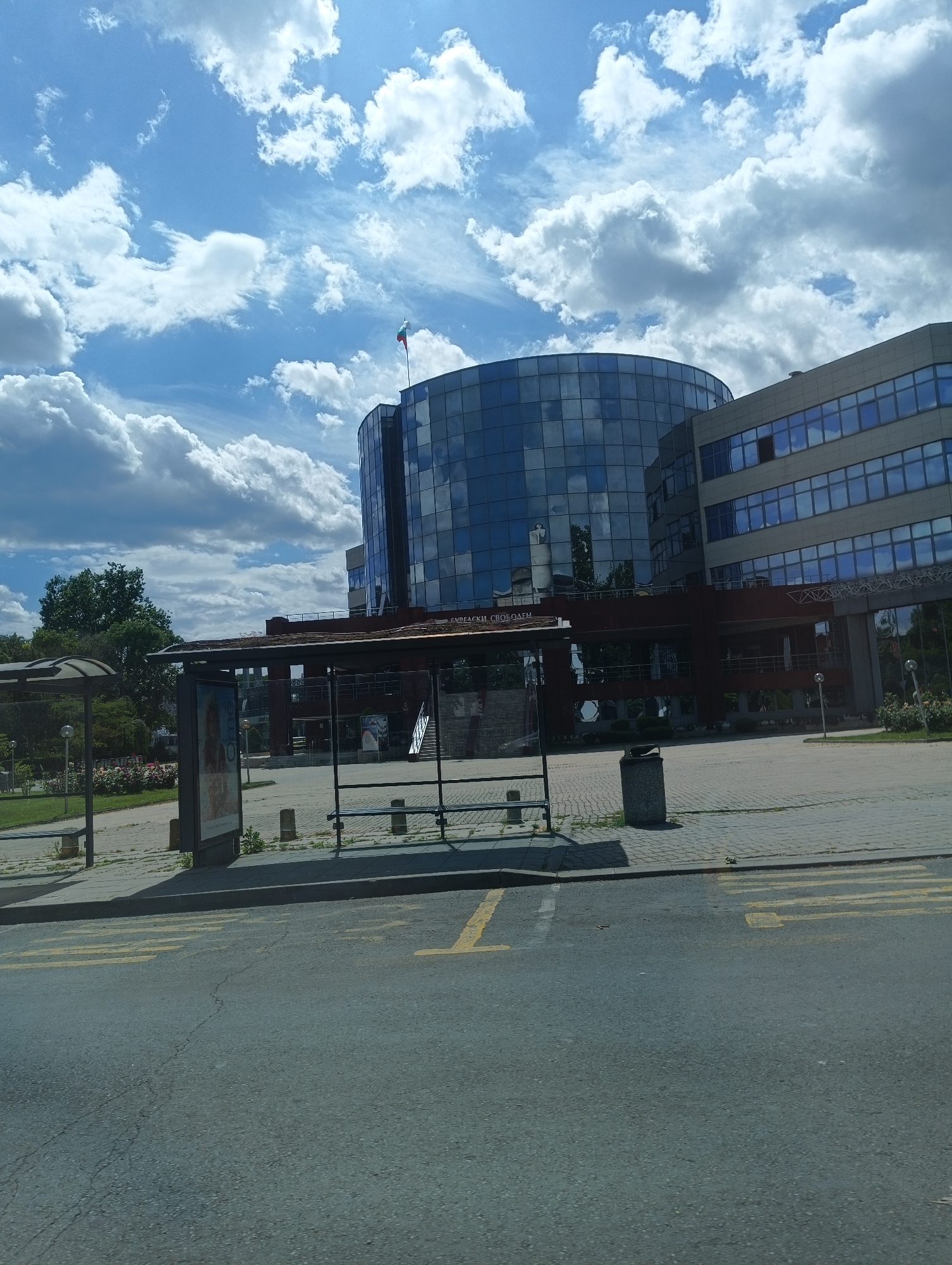 The image depicts a street scene in Burgas, Bulgaria. In the foreground, the asphalt road shows yellow road markings. A bus stop is visible, featuring a metal structure with seating. Behind the bus stop, a modern building complex dominates the view, characterized by a large, cylindrical glass facade reflecting the sky and clouds. A Bulgarian flag flies atop the cylindrical structure. The building adjoins a more conventional rectangular structure with rows of windows. Landscaping with trees and flowers is present around the buildings. The sky is partly cloudy with a bright, diffused light. The overall environment suggests an urban setting with modern architecture.