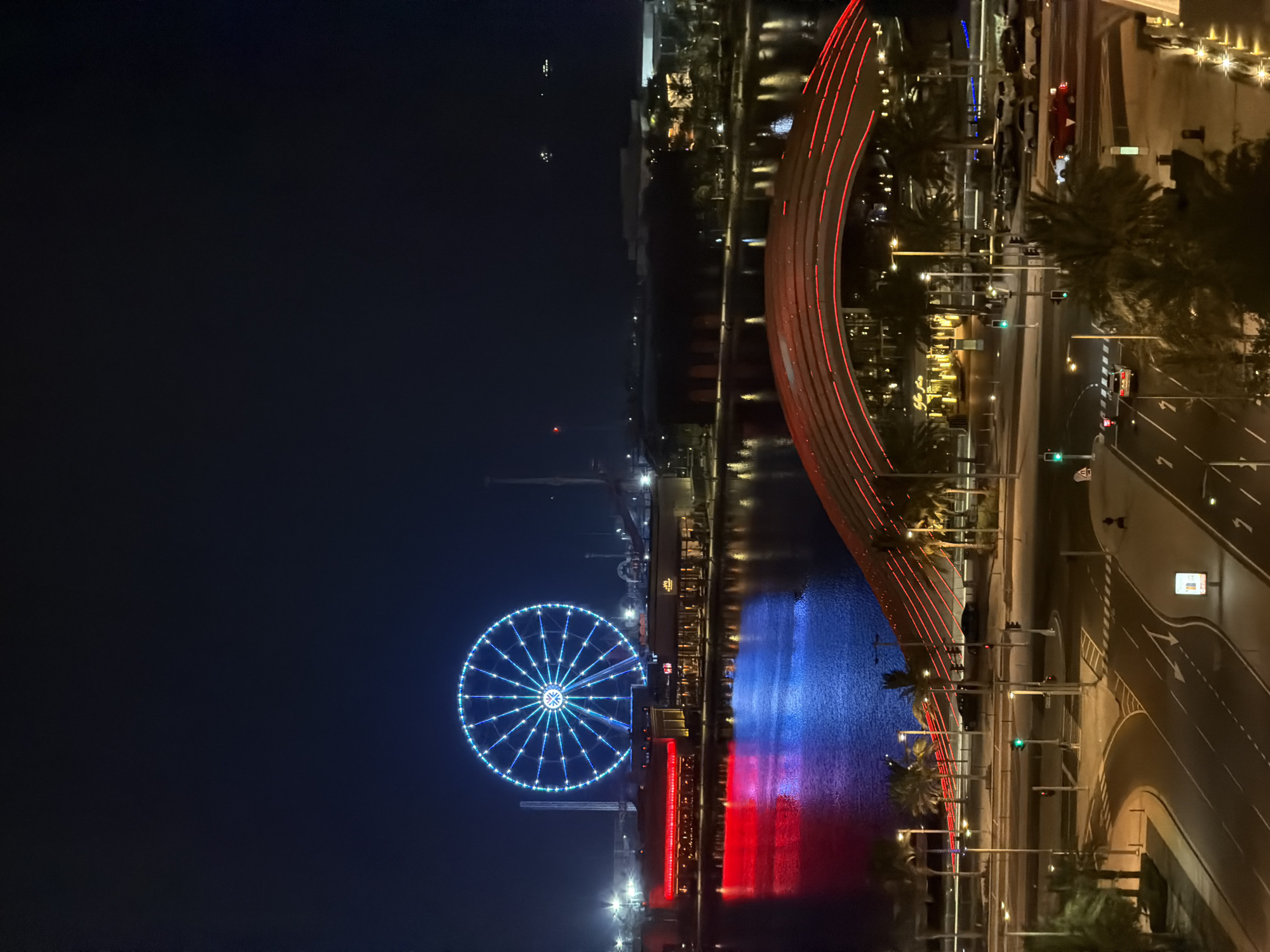 The image shows a nighttime cityscape with a large, illuminated Ferris wheel as the central focus. The Ferris wheel, with its blue and white lights, stands against a dark sky. Below it, a body of water reflects the colorful lights of the city, with red and blue hues dominating the reflections. Along the waterfront, buildings with red and yellow lights are visible, suggesting commercial or entertainment establishments.
In the foreground, a modern bridge with red illuminated railings arches over a street. The bridge's structure creates a dramatic visual element. Below the bridge, a multi-lane road is visible with streetlights and traffic signals. Several cars are present on the road, some parked and some in motion. Palm trees are scattered throughout the scene, adding to the ambiance. The overall impression is that of a vibrant urban area at night, likely a tourist or entertainment district. The Ferris wheel and the illuminated bridge suggest a lively atmosphere.