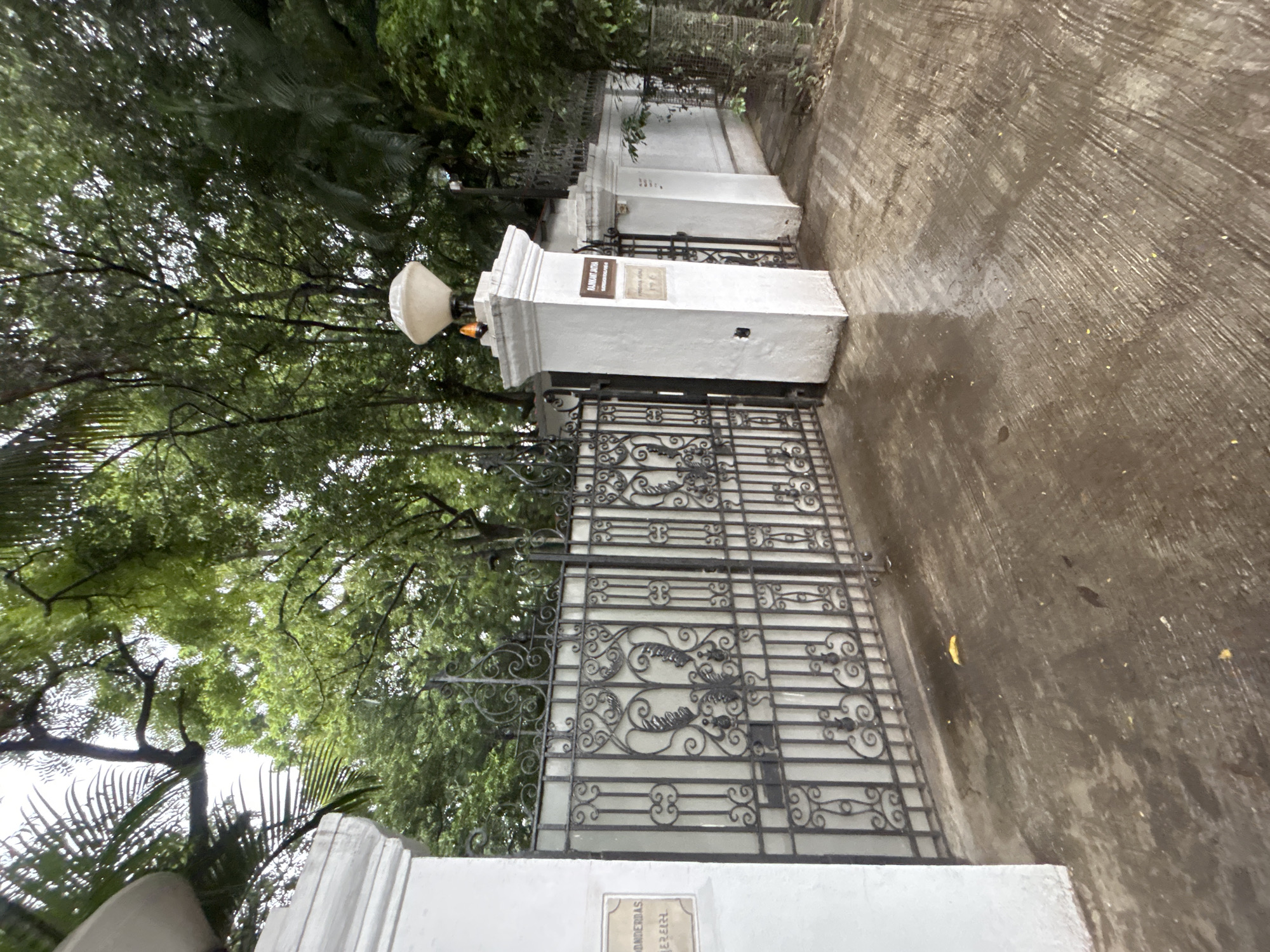 The image shows the entrance to a property at 12 Juhu Road, Pune, India. A tall white pillar with a decorative lamp on top stands to the right of a black wrought-iron gate. The gate is intricate, with swirling metalwork and decorative motifs. A plaque on the pillar reads "RAJNI KANT JHA." To the left of the gate, another white pillar has a smaller plaque with what appears to be an address. The ground in front of the gate is a wet, paved surface, suggesting it has recently rained. Lush green trees and foliage are visible behind the gate and pillars, suggesting a well-maintained garden or property. The overall impression is of a grand and established residence.