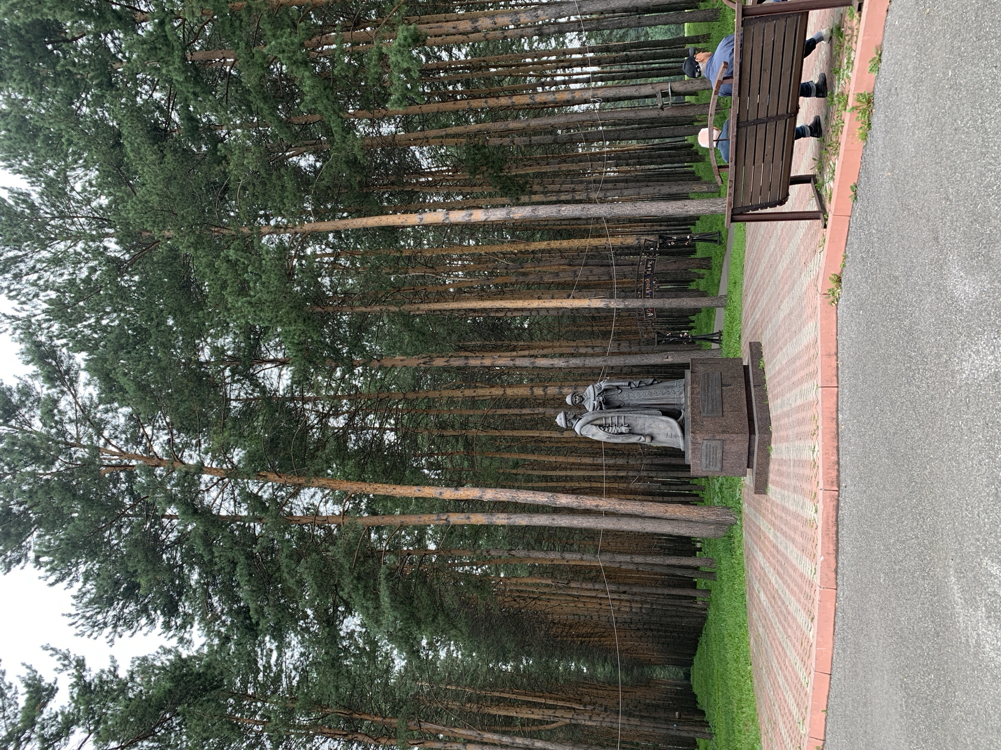 In Prokopyevsk, Russia, a statue of two figures stands at the entrance to a dense pine forest. The statue depicts two individuals in traditional attire, one appearing to be male and the other female, standing on a pedestal. The pedestal is inscribed with text, though it is not fully legible. The statue is located on a paved area with red and grey brickwork. To the right of the statue, two men are seated on a wooden bench, facing away from the camera. The forest is characterized by tall, slender pine trees with lush green foliage. The ground in front of the statue is covered with grass. In the background, a sign with Cyrillic lettering is visible, indicating it is the entrance to a park. The overall scene conveys a sense of history and nature, with the monument serving as a focal point within the park.