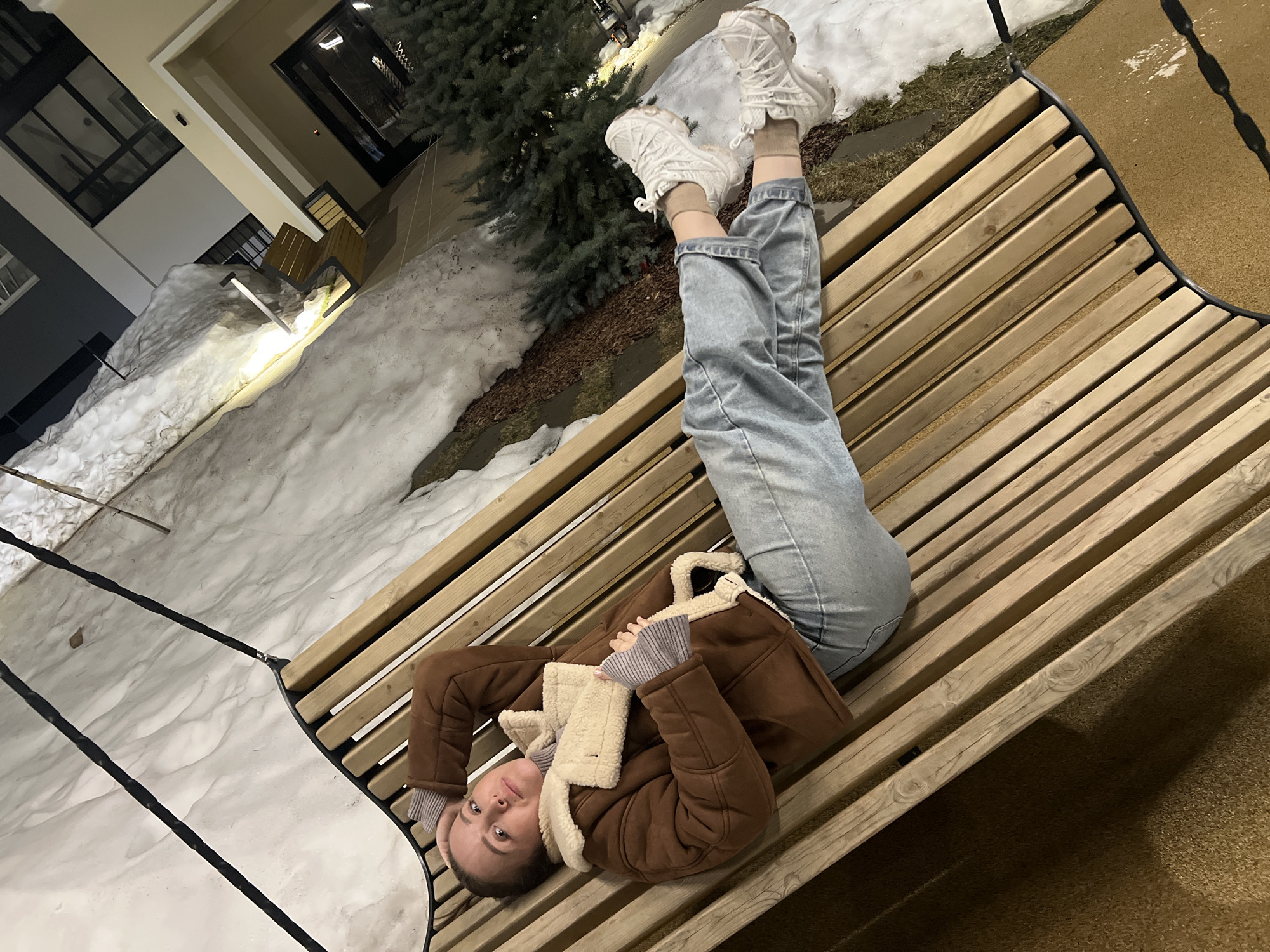 A female individual is depicted lying supine on a slatted wooden swing-bench in an outdoor amenity area, within an unknown urban or suburban location. The scene is illuminated by artificial lighting, indicating dusk or night conditions.
The subject, a woman with light brown hair and fair skin, wears a brown faux shearling jacket with cream-colored lining, a striped long-sleeve top, light blue denim jeans, white athletic shoes, and tan socks. Her left arm is bent with her hand positioned behind her head, while her right arm rests across her midsection. She is looking directly forward, towards the observer.
The swing-bench is constructed of light-colored wooden slats on a dark metal frame, suspended by dark ropes. The ground immediately beneath the swing consists of a dark tan granular surface with white spatter. Adjacent areas of the ground are partially covered with undisturbed snow and dark brown mulch, interspersed with evergreen shrubs. In the background, a modern, multi-story building with light-colored facades, dark-framed windows, and balconies is visible, partially lit. A vertical light fixture stands near the building's base in a snowy patch.