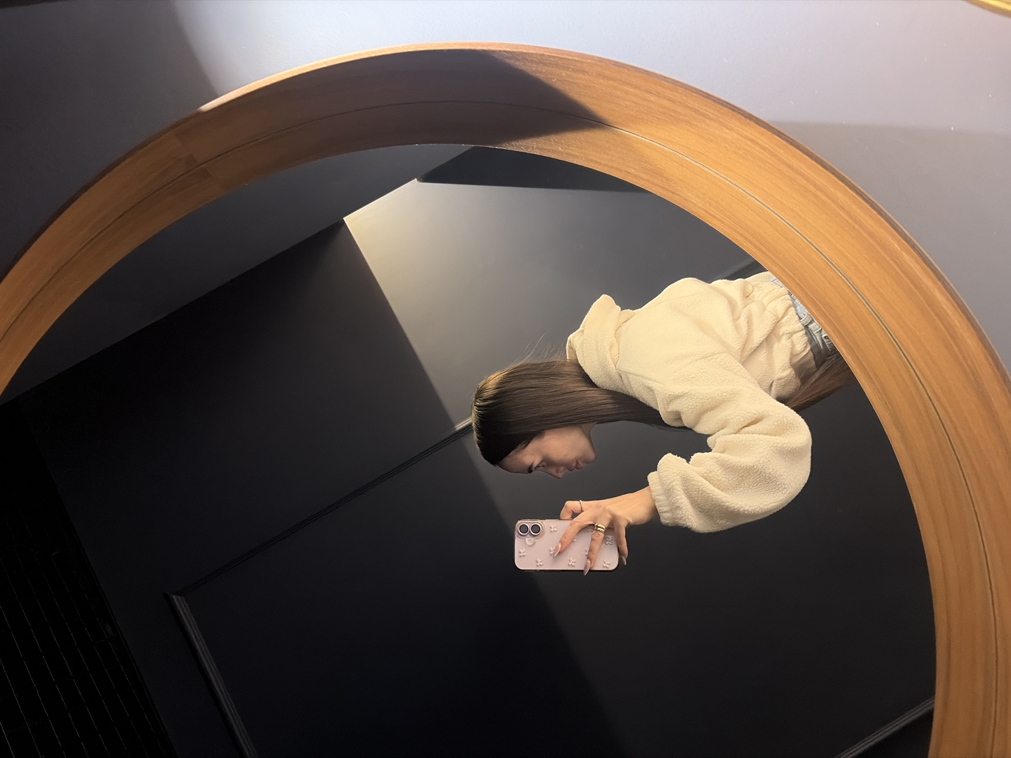 A woman is reflected in a large round mirror. The woman has long brown hair and wears a black headband, a light-colored fuzzy jacket, and jeans. She holds a pink smartphone in her hand. The background reflected in the mirror is a dark gray or black wall, possibly with panels or trim. The mirror's frame is made of light-colored wood. There is no information to suggest that this photo was taken in Saint Petersburg, Russia.