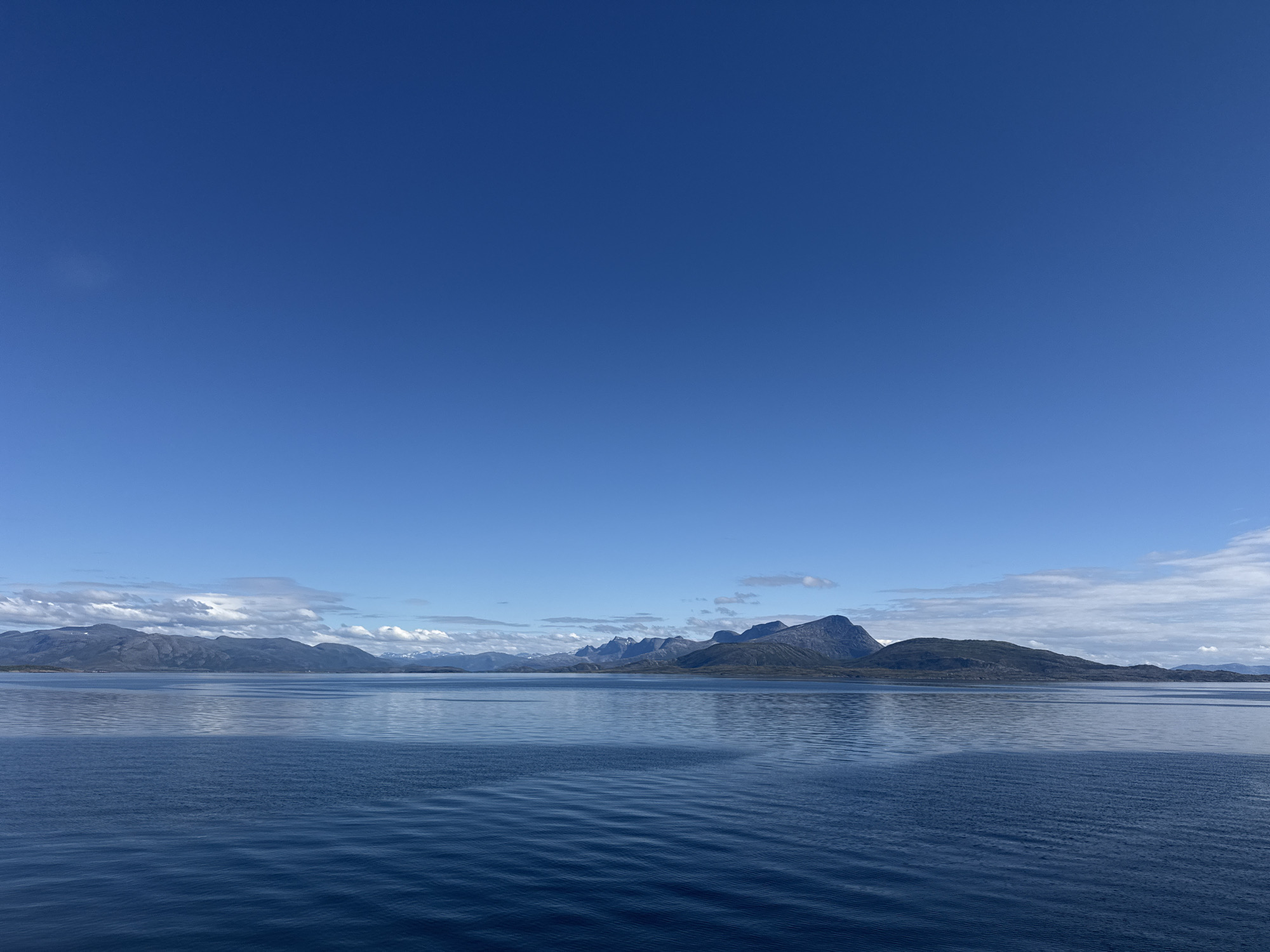 The image depicts a vast expanse of deep blue water under a clear, bright blue sky. In the distance, a rugged mountain range with some snow-capped peaks stretches across the horizon. The mountains appear to be covered in green vegetation in their lower slopes, and their peaks are sharp and imposing. The water is calm, with gentle ripples reflecting the colors of the sky and the distant landscape. The overall impression is one of serene natural beauty. This scene is likely located in the vicinity of Lødingen, Norway, a municipality known for its stunning coastal scenery and dramatic mountainous terrain.