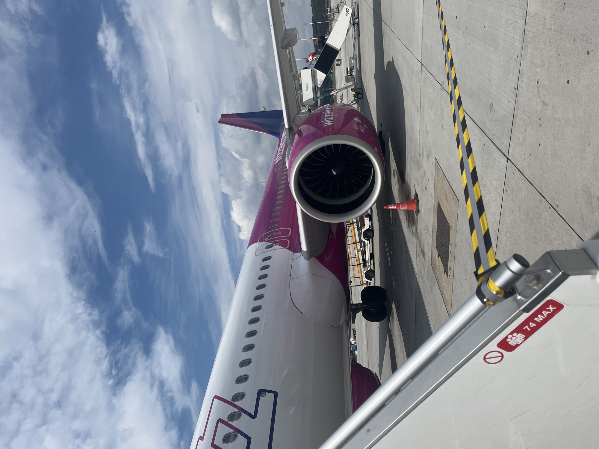 An airplane with Wizz Air livery is parked on the tarmac at Balice John Paul II International Airport near Krakow, Poland. The aircraft is primarily white with bright pink accents, including the tail fin and engine nacelles. A pink stripe runs along the fuselage, and the airline's logo is visible on the tail. The plane's engines are prominently featured. In the foreground, a mobile boarding stairs is positioned next to the aircraft. A red and white sign on the stairs indicates a maximum capacity of 74 people. The ground is concrete, marked with yellow and black striped hazard tape and other painted lines. Other airport vehicles and equipment are visible in the background, along with a red traffic cone. The sky is partly cloudy with blue sky visible between the clouds.