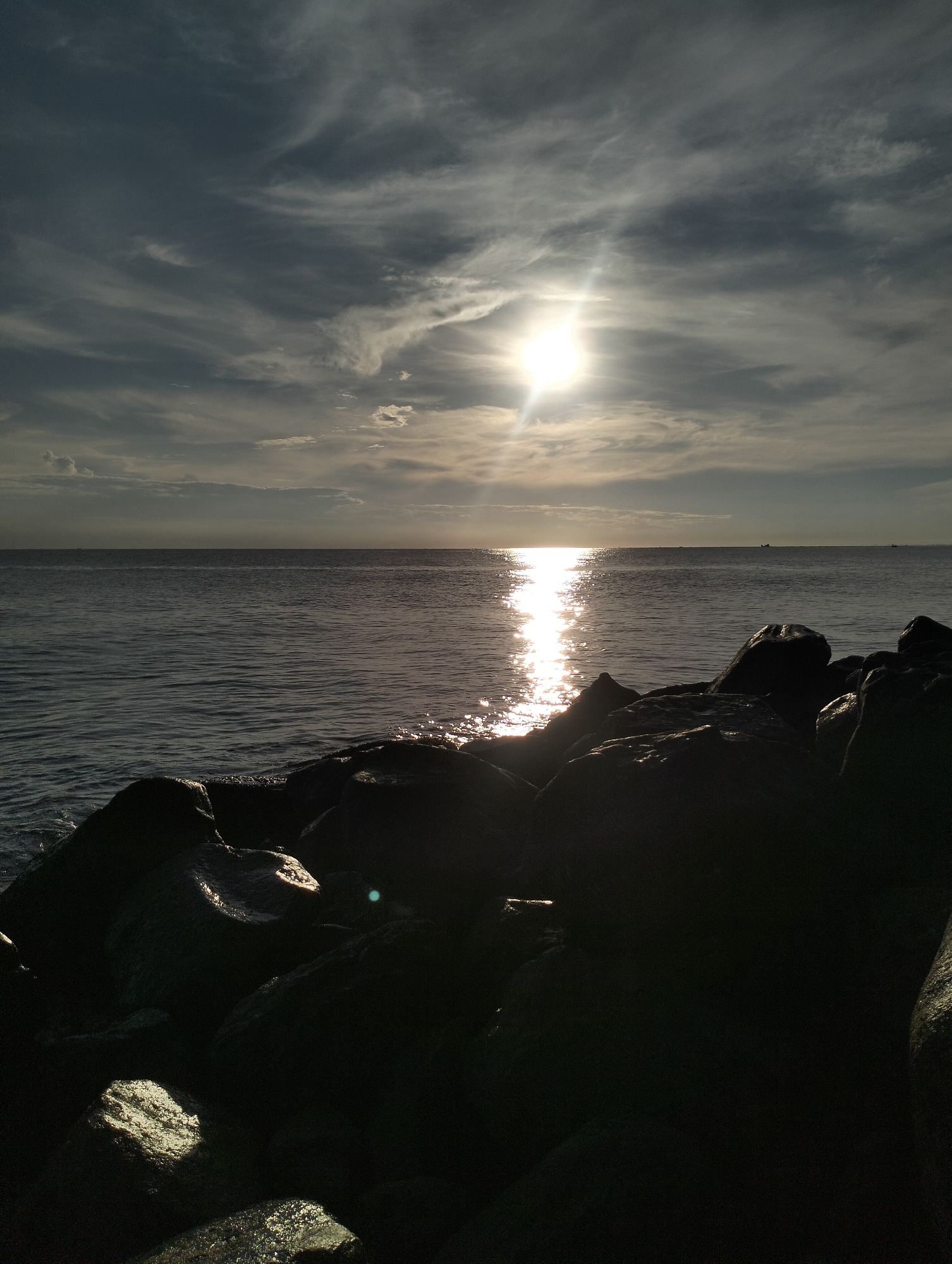 A landscape image shows the ocean, sky, and rocks. The sky is cloudy with the sun shining through the clouds. The sun's reflection shines down the ocean. In the foreground are dark, wet rocks. Lhokseumawe, Indonesia, is not discernible from the image.