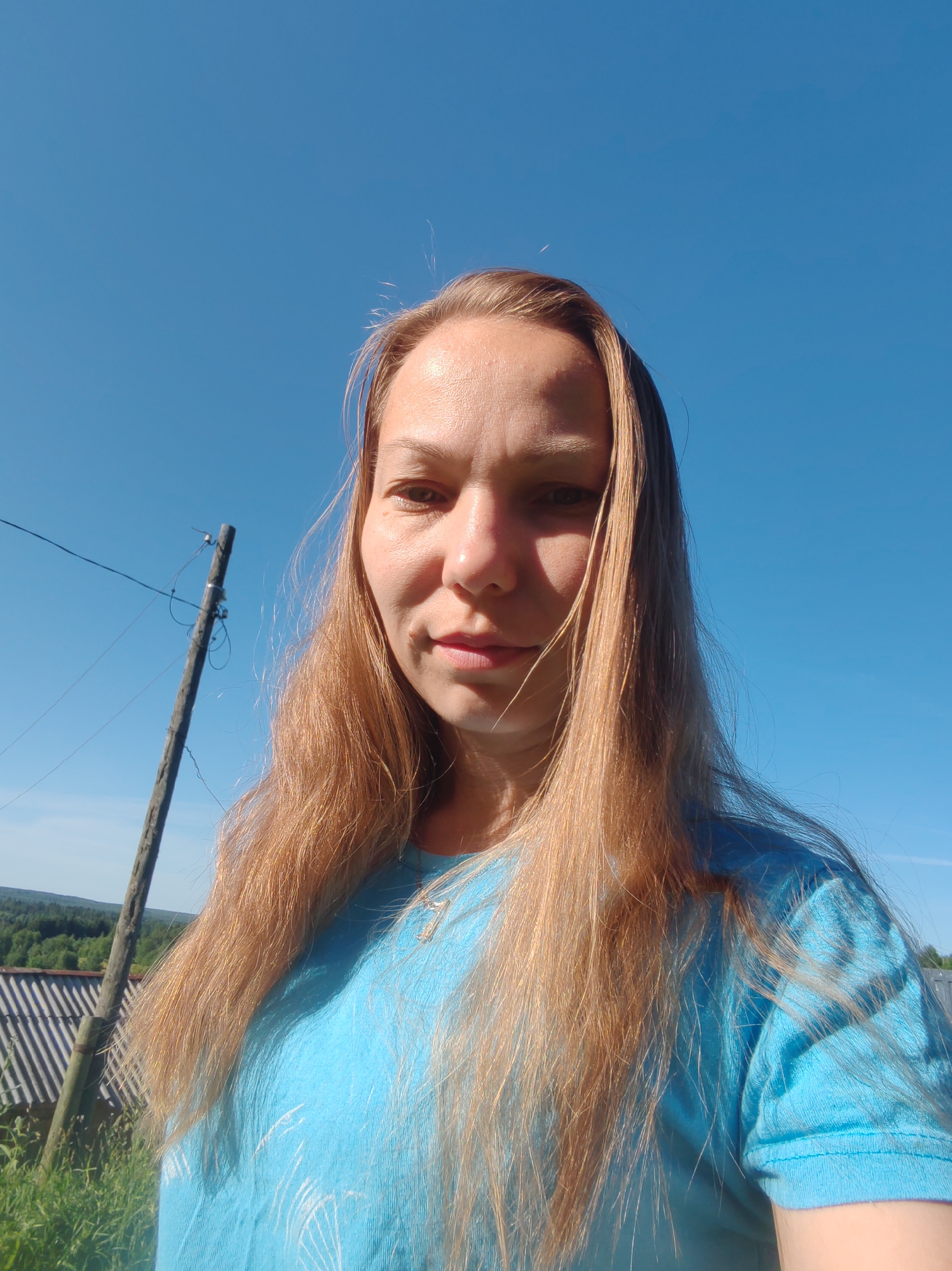A medium shot selfie captures a woman with long, blonde-brown hair against a clear blue sky. She is wearing a light blue t-shirt. Her face is turned slightly towards the camera, with a hint of a smile. Sunlight illuminates the right side of her face, casting a shadow on the left. Behind her, a wooden utility pole with wires is visible on the left side of the frame. To the left of the pole, a section of a rustic, grey roofed structure is visible, with green grass and trees in the background, suggesting a rural or suburban setting. The sky dominates the upper portion of the image. The overall impression is a casual, outdoor portrait on a sunny day. The location is identified as Chukhlem, Russia.