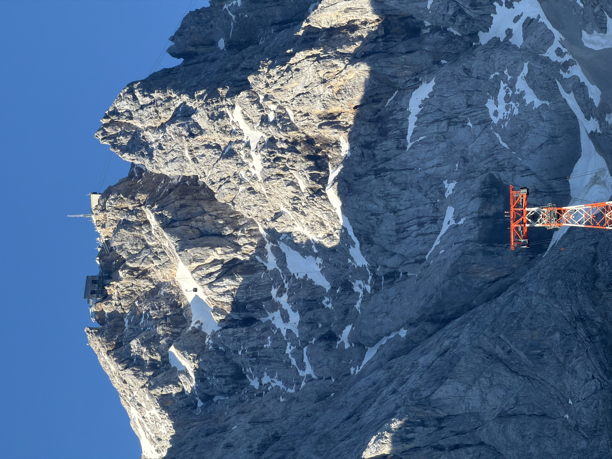 The image depicts the Zugspitze, Germany's highest peak, located near the municipality of Grainau. A cable car tower, painted in red and white, stands prominently in the foreground. Above it, the rugged, rocky slopes of the mountain rise towards the summit, with patches of snow scattered across the gray rock. At the very top, structures are visible, including buildings and what appears to be a weather station or observation deck. A small cable car is seen suspended on a line ascending the mountain, suggesting active transportation to the summit. The sky above is a clear, deep blue, indicating fair weather. The lighting suggests either early morning or late afternoon, with shadows cast across the mountainside.