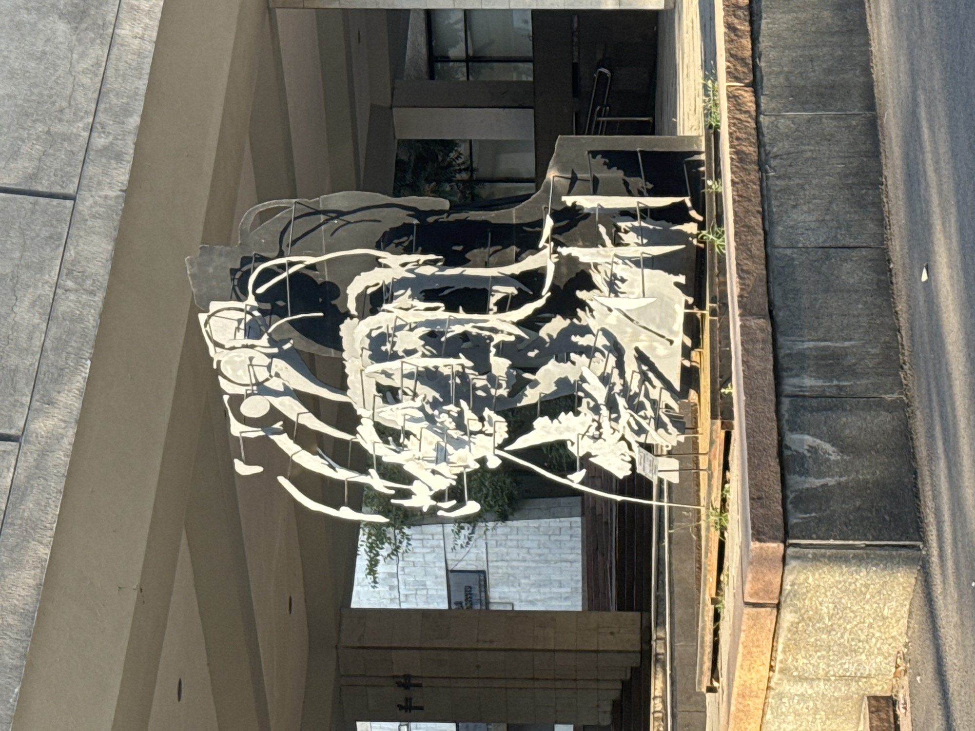 The image shows a large, abstract metal sculpture situated in front of a modern building with a colonnade. The sculpture is predominantly black and white, with an intricate, layered design that suggests human figures or faces. It rests on a raised, multi-tiered platform made of stone. Behind the sculpture, the building features a patterned facade and large windows. The setting appears to be outdoors, possibly a public space or plaza.
The sculpture's location is identified as the Monument to Manas, situated in Bishkek City, Kyrgyzstan. This landmark commemorates Manas, the epic hero of Kyrgyz folklore. The building behind the sculpture is likely part of the administrative or cultural complex surrounding the monument. The overall scene captures a moment of stillness, with the sculpture as the central focus, bathed in the natural light of what appears to be late afternoon or early morning, given the shadows. There are no people visible in the frame.