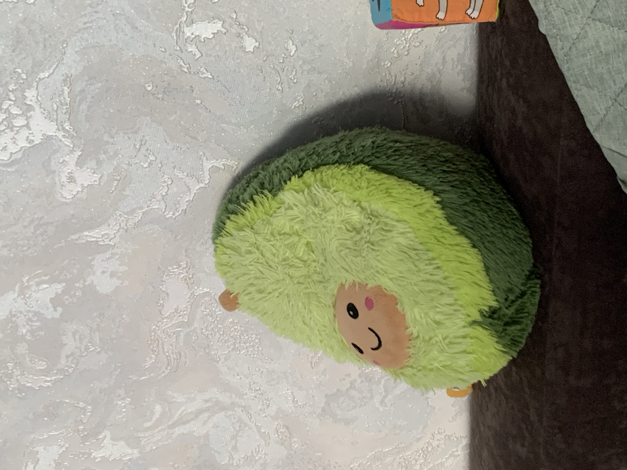 A plush avocado toy is the central subject, lying on a textured, light-colored wall. The avocado is depicted in a cute, cartoonish style with a round face featuring black eyes, a smiling mouth, and two pink circles on its cheeks. It has a fuzzy texture and is divided into segments of light and dark green, resembling the fruit's interior and skin. A small, orange-brown detail, possibly representing the avocado pit or a limb, is visible on the left side, and another similar detail is on the bottom right. The background is a light-colored, textured wall, possibly wallpaper, with a raised, abstract pattern. To the right of the avocado, a dark, rich brown surface suggests furniture, perhaps a sofa or a cushion. Above the avocado and slightly to the right, a colorful object, likely another toy or a decorative item, is partially visible. It appears to be a flat, rectangular object with orange and white colors, featuring cartoonish white legs. The lighting appears to be artificial, casting a soft shadow behind the avocado, suggesting an indoor setting. The overall scene is domestic and casual, likely a child's room or a living space. There is no visible text in the image. The image's orientation is sideways.