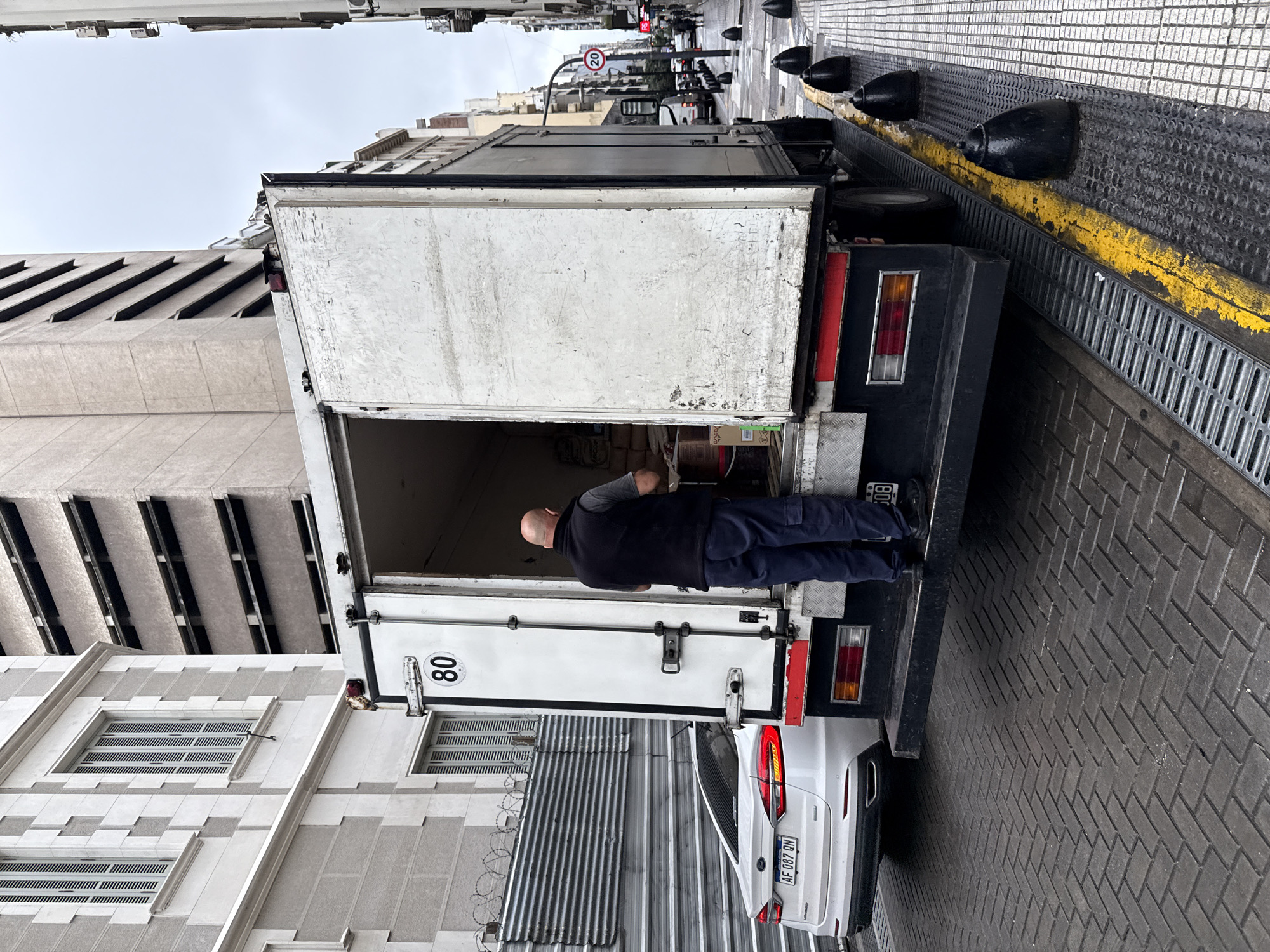 This media file depicts an urban street scene in Buenos Aires, Argentina, under an overcast sky. The central focus is a large white delivery truck, parked with its rear door open. A bald man, dressed in a dark vest, long-sleeved shirt, and dark pants, is standing at the back of the truck, actively engaged in loading or unloading items from its cargo area.
The truck is positioned alongside a paved area featuring a dark, textured surface with a prominent yellow stripe and a metal drain grate running parallel to the street. Several black bollards are spaced along this paved strip. Behind the truck, a white car, identified by its Argentinian license plate "AF 087 QN," is parked.
The surrounding environment includes architectural details that suggest a city setting. To the immediate left of the truck, a building with a textured facade of light and dark grey panels and multiple window openings is visible. Below this, a corrugated metal fence topped with barbed wire adds an urban detail. In the background, down the street, additional buildings line the road, and a traffic sign displaying "20" (indicating a speed limit) can be seen. Other vehicles are also present further down the street. The scene captures a typical daytime commercial activity, likely a delivery, in a bustling urban context.