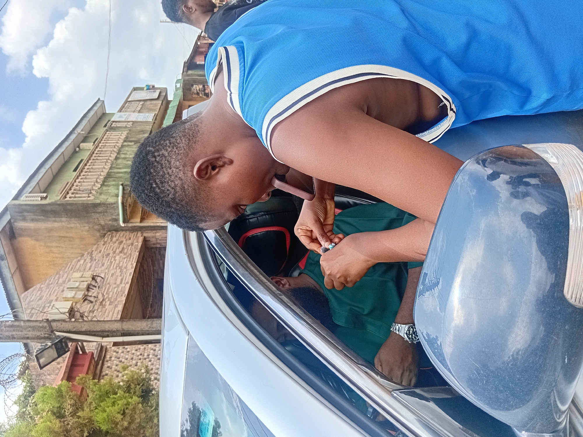 A photograph shows a man in a blue polo shirt leaning into the open window of a silver car. The man is holding a small object in his hands, and he appears to be interacting with someone inside the car, who is wearing a green shirt. The reflection of a building is visible in the car's window. The presence of dense vegetation, and a building with barbed wire on its walls indicates an urban setting within the city of Ikorodu, Nigeria.