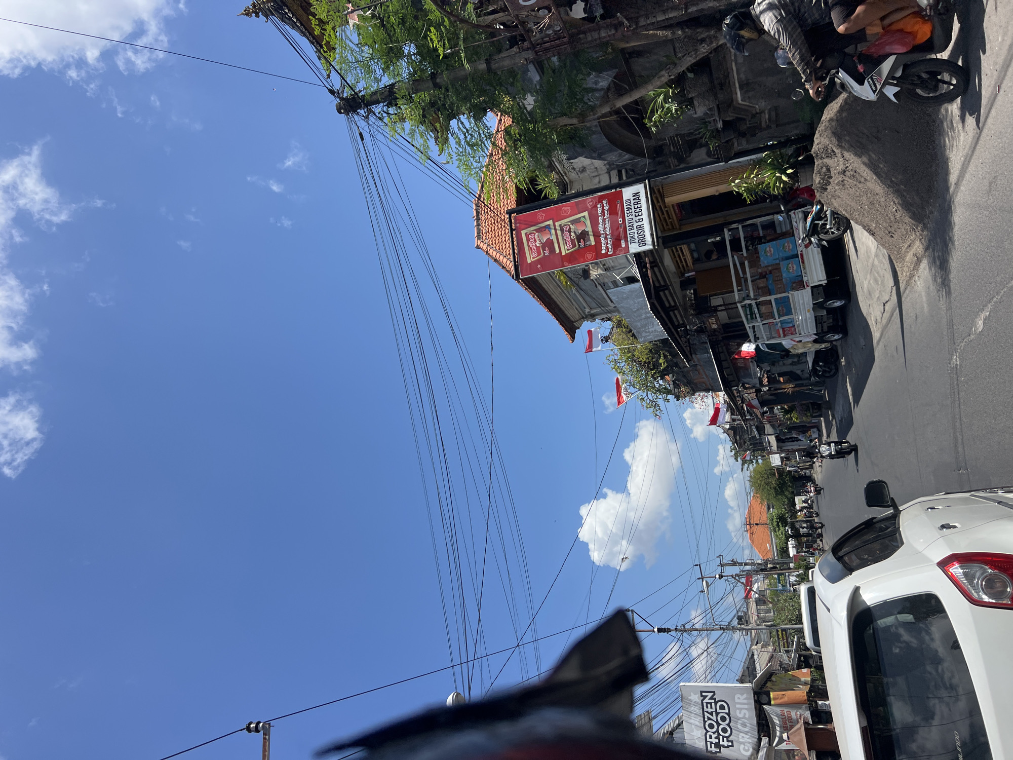 The image depicts a street scene in Denpasar, Indonesia, under a bright blue sky with scattered clouds. The road is paved and appears to be a busy thoroughfare with vehicles and pedestrians. On the right side of the street, a pile of sand is prominent, with a motorcycle rider in the foreground, facing away from the camera, wearing a helmet and casual clothing. Behind the sand pile, a white truck is parked, loaded with blue boxes. Buildings line the street, with traditional Balinese architecture visible in some structures, including tiled roofs and ornate carvings. Several red and white flags, possibly Indonesian flags, are displayed on poles and attached to buildings. Signs in Indonesian are present, with one advertising "FROZEN FOOD GROSIR". The overall impression is of a bustling urban environment in Indonesia.