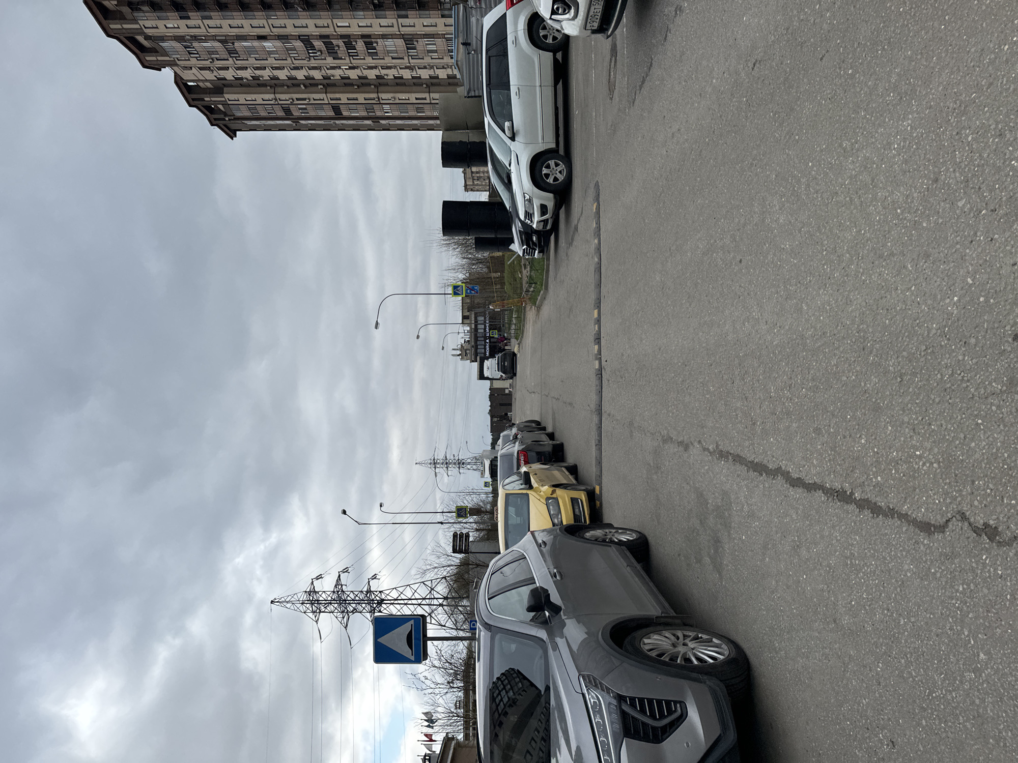 The image depicts a street scene in Odintsovo, Russia, characterized by a row of parked cars along the right side of the road. Dominating the foreground is a grey SUV, positioned at an angle, partially obstructing the view of the street. Behind the SUV, several other cars are parked, including a yellow sedan, and vehicles of various colors that extend into the distance. The street surface is asphalt, showing signs of wear with visible cracks. On the left side of the road, an electrical transmission tower is visible, standing next to a road sign displaying a warning symbol. Beyond the sign, there is greenery and other structures. A large building looms on the top left of the image. The sky is overcast with heavy clouds, indicating a cloudy day. There are no visible people in the scene.