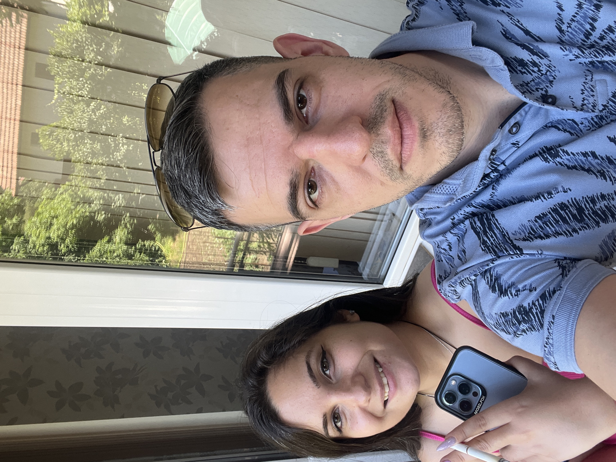 A man and a woman are taking a selfie outdoors, near a window with a view of trees and a building. The woman is on the left, smiling and holding a smartphone and a cigarette. She is wearing a pink top and has long, dark hair. The man is on the right, looking at the camera with a neutral expression. He is wearing a blue polo shirt and has short, graying hair and stubble. The background shows a patterned wallpaper on the left and a window with greenery and building reflections on the right. The lighting suggests it is daytime. The location is not specified in the image.