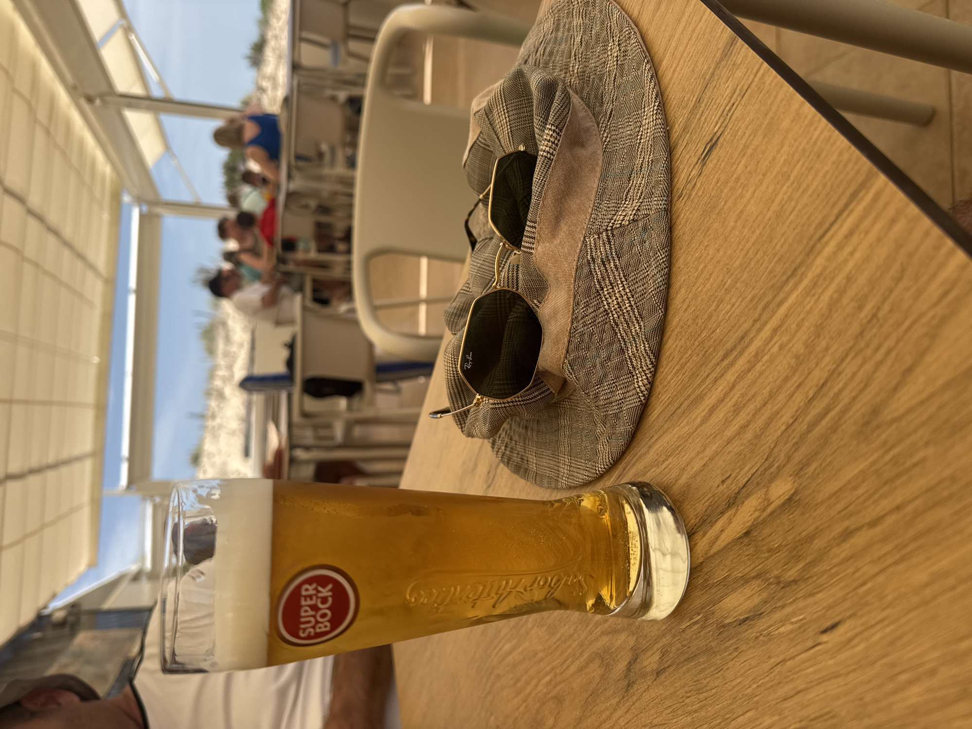The image captures a moment in an outdoor dining setting, likely a café or restaurant, in Unknown, Unknown. The primary focus is on a table displaying a glass of Super Bock beer, a patterned hat, and a pair of Ray-Ban sunglasses. The table has a light brown wood-grain surface. The beer glass is tall and slender, partially filled with a golden liquid, with the Super Bock logo prominently visible. The hat, resting beside the beer, features a grey and white checkered pattern. The sunglasses, placed on top of the hat, have dark lenses and gold frames.
In the background, blurred figures of people are seated at other tables, suggesting a populated establishment. A partial view of the building's structure includes white awnings. The scene has a bright, daytime ambiance.