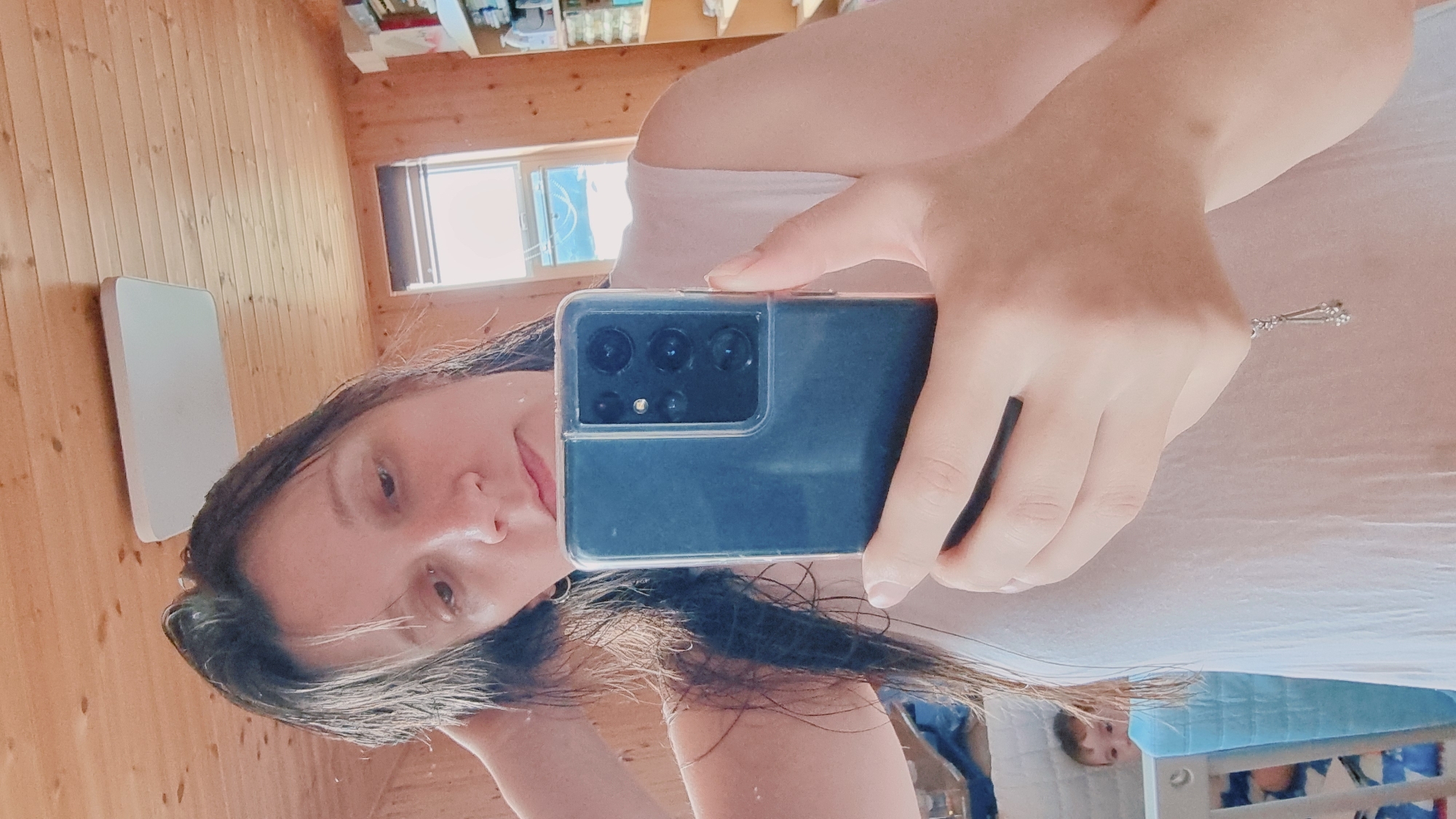 A woman is holding a cell phone and taking a selfie in a room with a wooden wall. The woman has long, dark hair and is wearing a light-colored top. The phone is black, and the camera lenses are visible. A window is in the background, and there is a white object on the wall. In the lower right corner of the image, a child can be seen in a crib. The location is likely indoors at a home or residence, in the city of Dunnae-myeon, South Korea.