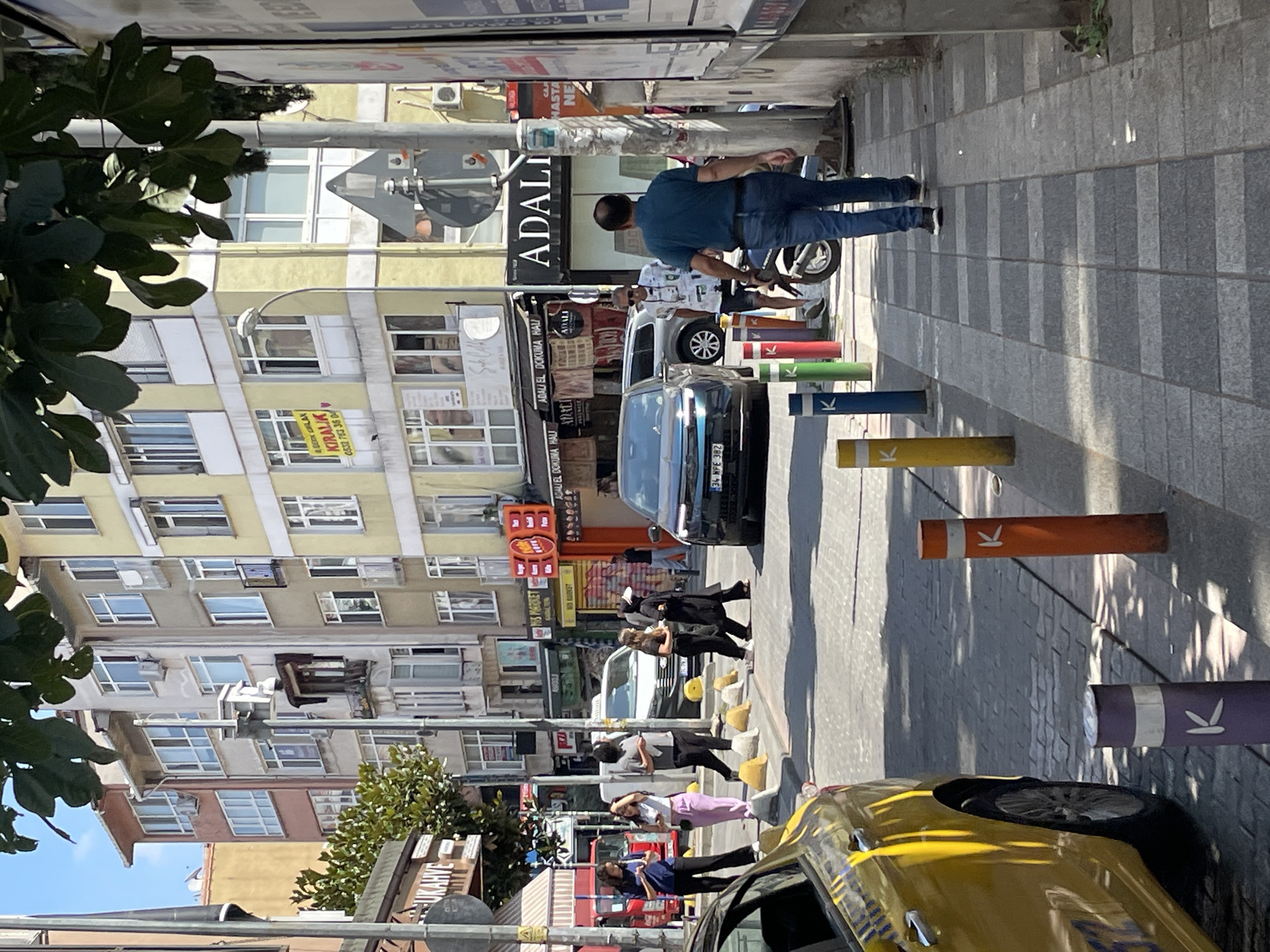 The image depicts a street scene in Kadıköy, Turkey. In the foreground, a yellow taxi is parked on the left, partially visible. A man in a blue polo shirt and jeans walks away from the camera on a paved sidewalk. Colorful bollards line the edge of the sidewalk, with the letter "K" visible on some of them. In the middle ground, several people are crossing the street, including a group of women in casual attire and another person in a dark abaya and headscarf. A black car and a white van are stopped on the street. Behind the street, multi-story buildings with numerous windows line the thoroughfare. Some buildings display signs for businesses, including "ADALI EL DOKUMA HALI" and "BİZ MARKET". The overall environment appears to be an urban commercial area on a sunny day.