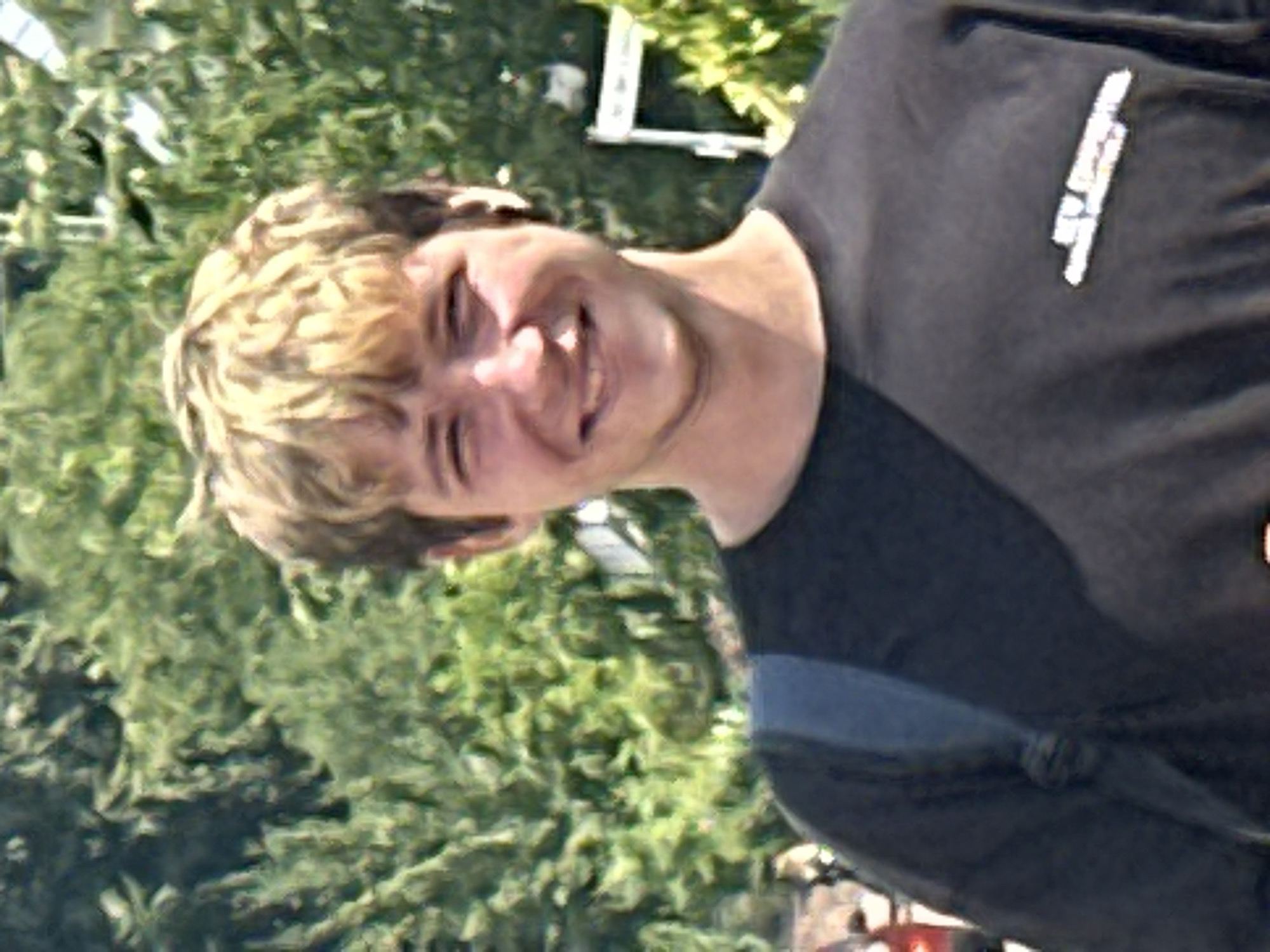 A young Caucasian male with blonde hair is standing in front of a lush green backdrop of trees. He is wearing a dark t-shirt with a graphic on the left chest. A blue strap of a bag is visible across his shoulder. The man is smiling at the camera. The image is likely taken outdoors in a park or natural setting. There is no discernible information indicating the location in the city of Günzburg, Germany.