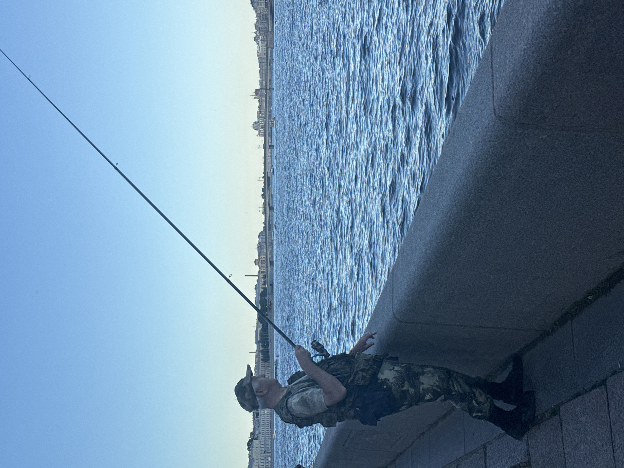A man is fishing on the embankment of the Neva River in Saint Petersburg, Russia. He is wearing a camouflage patterned baseball cap, a camouflage vest over a t-shirt, and camouflage pants. He is holding a fishing rod with his right hand and his left hand is resting on the stone embankment. The water is choppy, with a cityscape visible in the background, including buildings, bridges, and a church dome. The sky is clear and blue, suggesting it is daytime. The embankment is made of large, rough-hewn stones, and a paved walkway is visible to the left of the man.