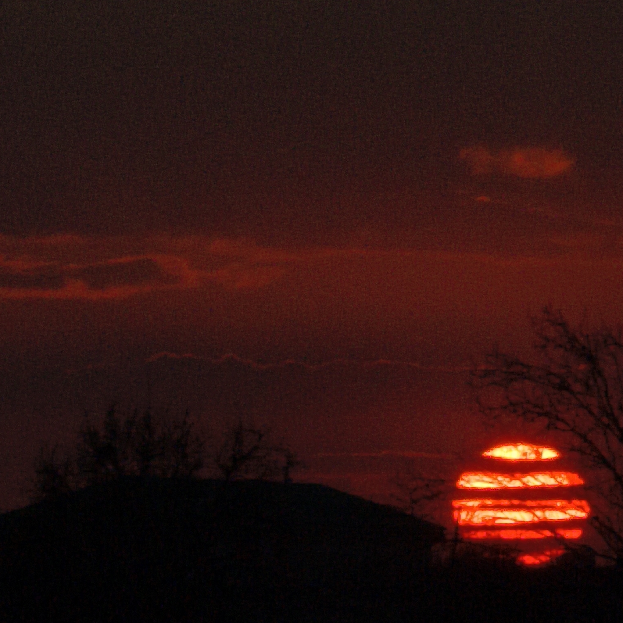 A dark and fiery sunset scene is captured, with the sun appearing as a horizontally striped, vibrant red orb low on the horizon. The silhouette of a tree with bare branches is visible on the right side of the frame, partially obscuring the sun. A darker silhouette of what appears to be a hill or a structure with sparse vegetation is present in the lower-left portion of the image. The sky exhibits a gradient of dark red and orange hues, suggesting atmospheric conditions conducive to a vivid sunset. Based on the known photograph details, the location is Hrodna, Belarus. The image suggests a natural scene with trees and a possibly elevated ground feature against the backdrop of an intense sunset.