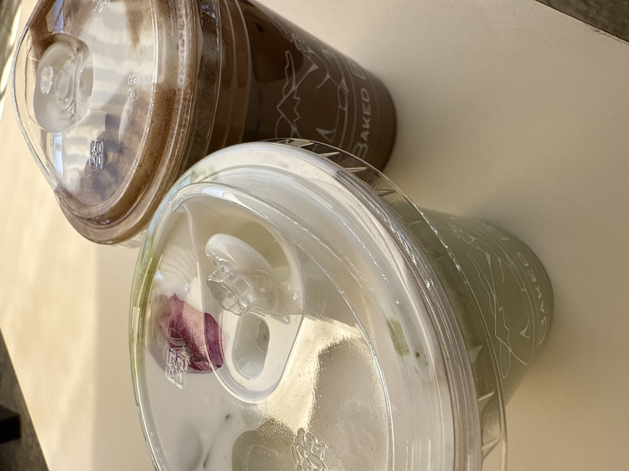 Two clear plastic cups containing cold beverages are displayed on a light beige tabletop in San Bernardino Tlaxcalancingo, Mexico. The cup in the foreground contains a light green liquid, consistent with a matcha beverage, topped with a layer of white foam. A small, dried purple rose petal rests on the foam, visible through the clear plastic dome lid, which features a flip-top sipping opening. The side of this cup bears white, stylized lettering, including the partial word "BAKED". Positioned behind and to the right is a second clear plastic cup containing a dark brown liquid, likely an iced coffee or chocolate drink, also topped with a lighter foam layer. This cup also has a clear plastic dome lid with a sipping opening. Its side displays white graphics, including a silhouette of mountain peaks and the complete word "BAKED". No individuals or other objects are visible in the scene.