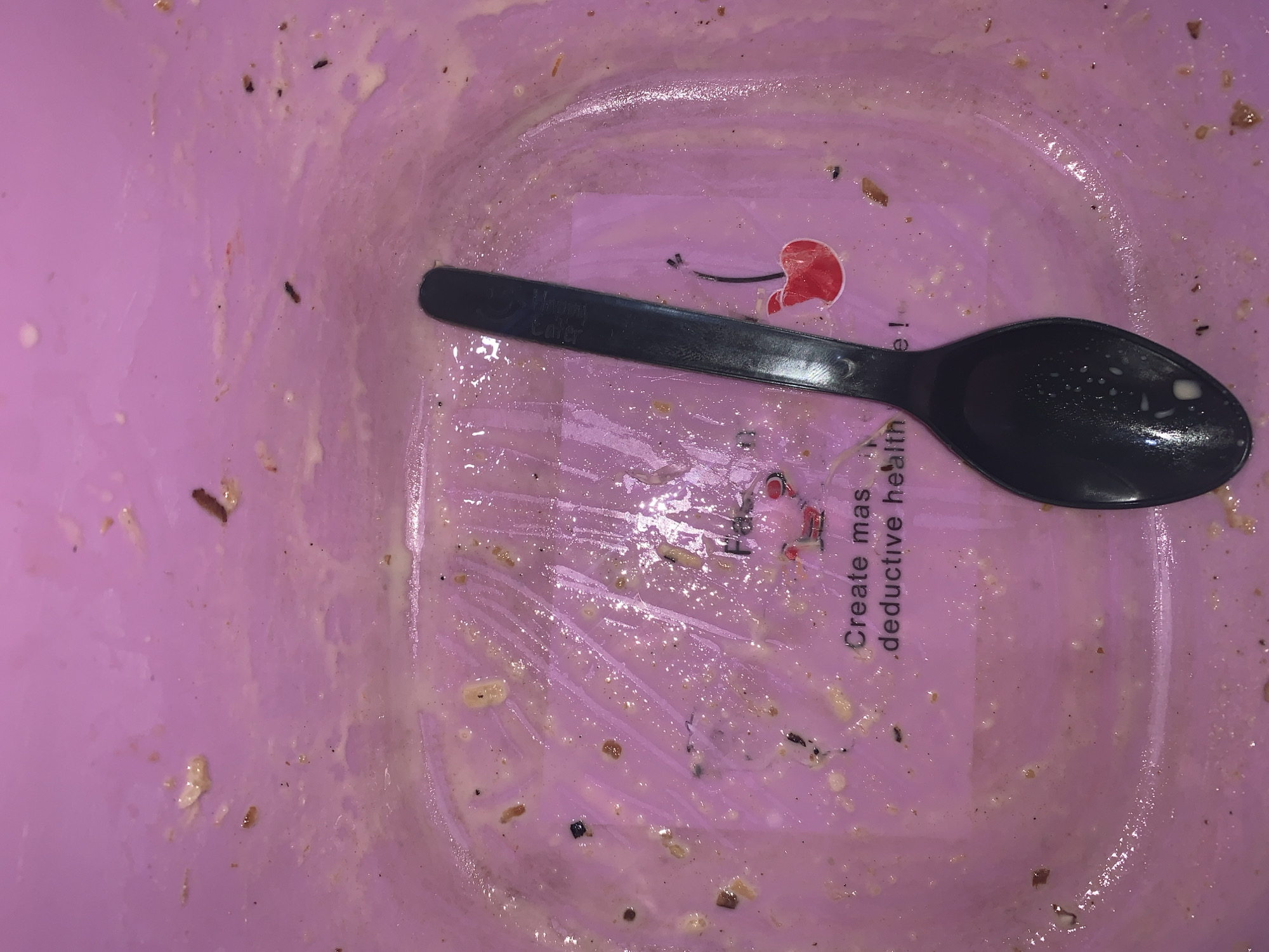 The image displays the interior of a purple plastic bowl, largely emptied of its contents but still showing significant residue of a creamy, light-colored food or liquid. Streaks and smears cover the inner surface, indicating the food has been mostly consumed, leaving behind scattered darker specks that suggest ingredients like spices or grains. A black plastic spoon rests horizontally across the bowl's bottom, also bearing traces of the food.
Faint branding and text are visible beneath the food residue. On the handle of the spoon, the words "Happy Eater" are subtly discernible. The bowl itself features a red graphic resembling a cherry with a stem. Adjacent to this graphic, partially obscured text reads "Create mas," and below it, positioned vertically, the phrase "deductive health" is visible. The scene captures the aftermath of a meal or snack, likely a creamy dish such as yogurt, soup, or a smoothie. No specific time of day or location cues for Rafin Par, Nigeria, are identifiable beyond the general setting of a consumed meal.