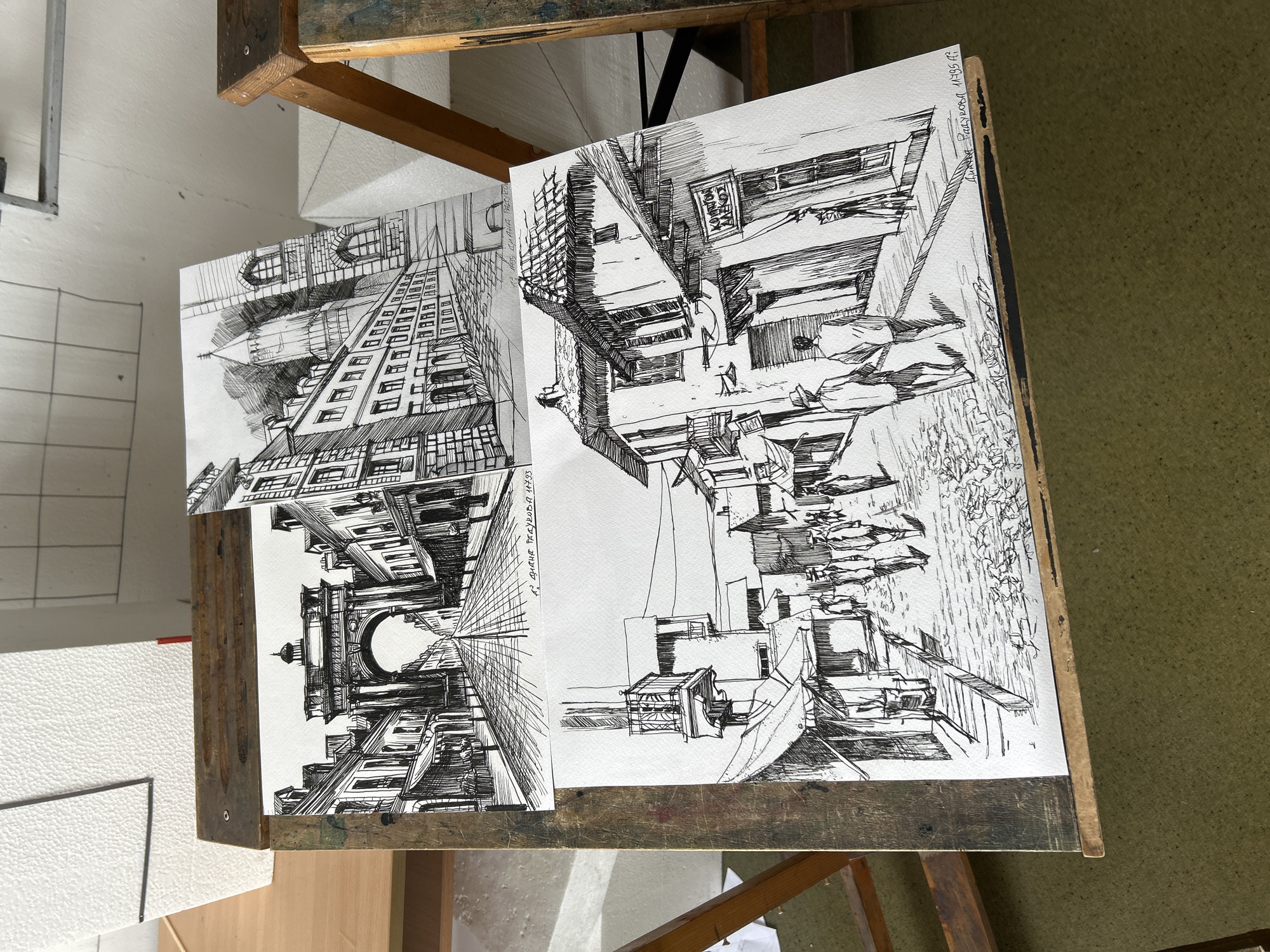The image shows two black and white architectural drawings on a wooden easel. The drawing on the left depicts a street scene with buildings receding into the distance, converging at a vanishing point, topped with a tall arch. The building on the right appears to be an urban environment that includes a storefront with an awning, outdoor seating, trees, and walking pedestrians. The perspective of each drawing is a single-point view. Both drawings feature buildings and pedestrian activities. Based on the style and subject matter, these appear to be pen and ink architectural sketches. The drawing on the left shows a date: “27 ДИЯН РЪЖДОВА 1983". The drawing on the right shows a date: "27 ДИЯН РЪЖДОВА 11:45 1983". Without additional information, I cannot specify the exact locations in Sofia.