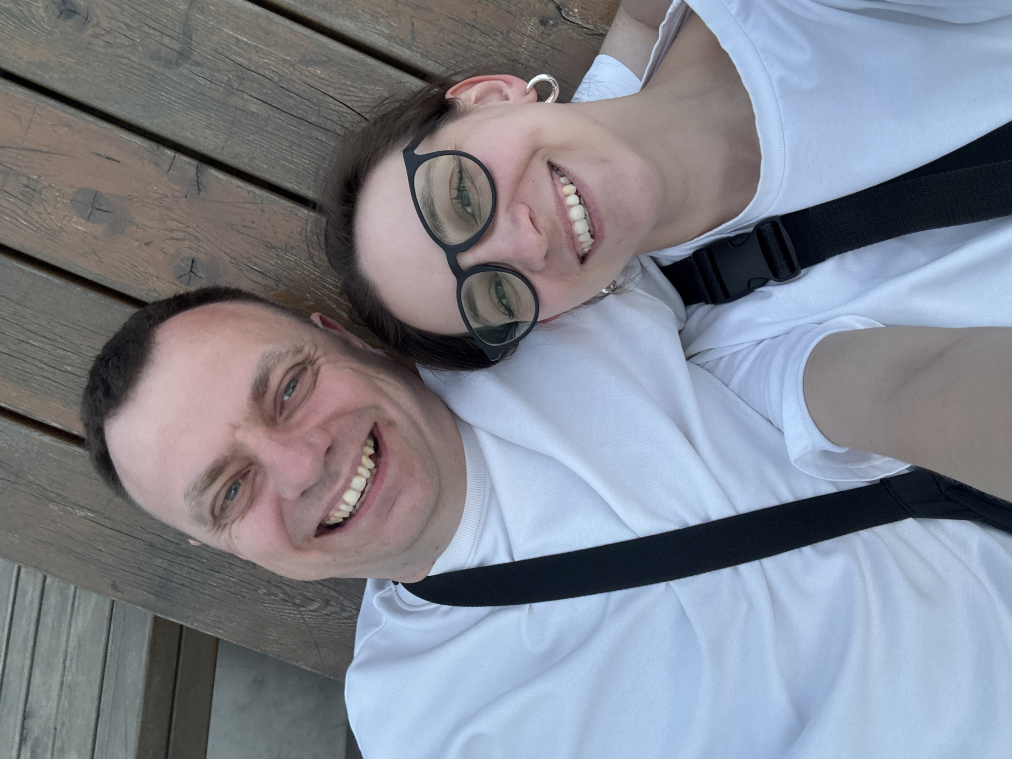 A selfie taken by a man and a woman lying on wooden planks. The man has short dark hair and is wearing a white shirt. The woman has her head resting on the man's shoulder. She has dark hair pulled back, wears glasses, and is also wearing a white shirt. A black strap is visible across both of their chests. The location is likely a public area, given the wooden planks, possibly in the city of Nizhny Novgorod, Russia.