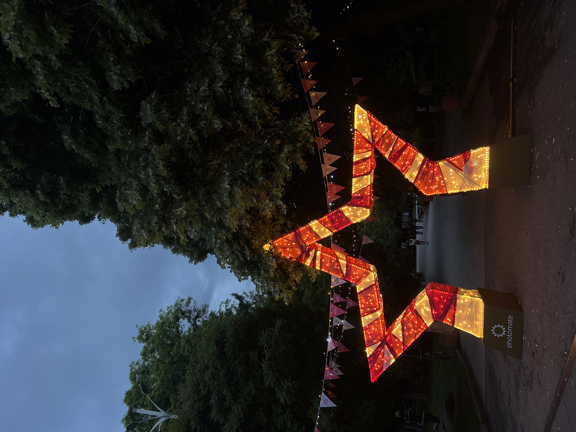 A brightly illuminated, oversized, red and white zig-zag shaped arrow stands in a park in Plovdiv, Bulgaria, likely for a festive event or advertisement. The arrow is mounted on boxy, dark green bases with "photomate" branding visible on one. Festive banners with triangular flags are strung nearby. A large tree with dense green foliage is visible in the background, along with a grey sky. The ground appears to be a combination of paving and grass. In the distance, several people are faintly visible walking. The scene is captured at dusk or night.