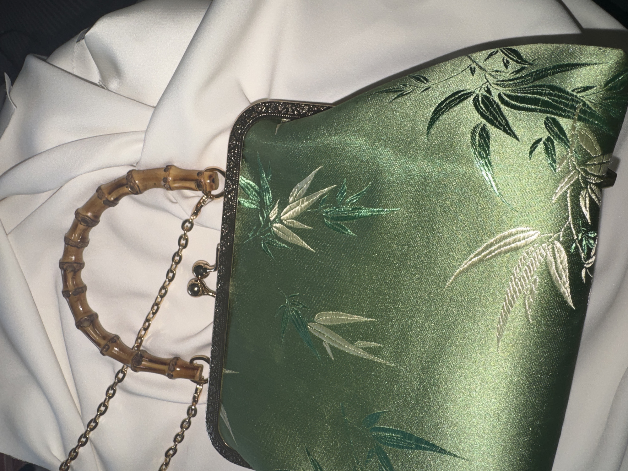 A close-up, high-angle shot focuses on a green satin handbag with a bamboo handle and a gold chain. The handbag is adorned with an embroidered bamboo leaf pattern in shades of green and gold. The bag is placed on a draped white fabric, possibly a tablecloth or garment.