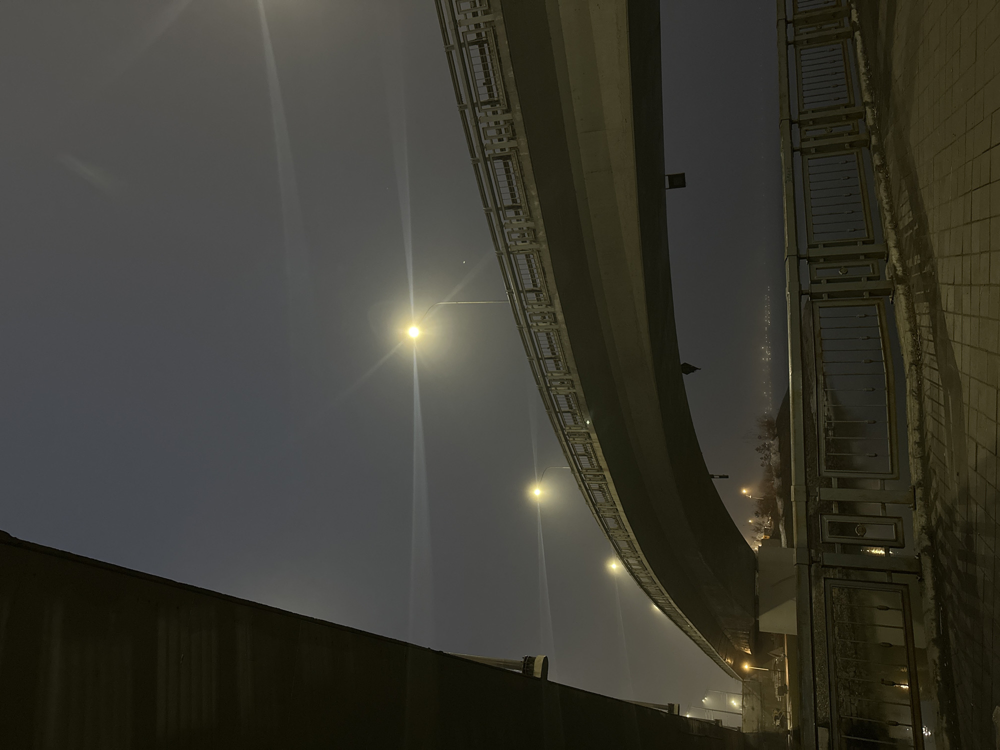 The image shows an urban night scene, likely in Kazan, Russia, featuring an elevated highway curving overhead. The sky is obscured by fog or haze, illuminated by streetlights that cast bright vertical beams. Below the highway, there's a low wall on the left and a metal railing or fence on the right, suggesting a pedestrian walkway or sidewalk. The ground is barely visible, but seems wet or damp, reflecting some light. The overall atmosphere is dim and slightly obscured.