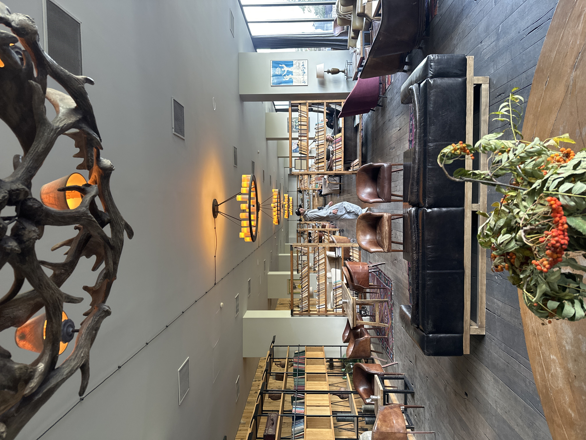 The image depicts a spacious, modern lounge or library area with rustic design elements, located in Stepantsminda, Georgia. The room features high ceilings with a unique antler chandelier and several modern, ring-shaped chandeliers with yellow lights. The flooring is made of dark, wide wooden planks.
The environment is furnished with a variety of seating options, including several distressed leather armchairs, a large black leather sectional sofa, and plush velvet chairs in shades of purple and brown. Wooden bookshelves line the walls, filled with books and decorative items. Several tables are scattered throughout the space, including small side tables and larger dining tables. A patterned rug is placed on the floor in the central seating area.
In the background, a person in a light-colored tracksuit walks through the room, and another person is visible sitting at a table. Large windows line one side of the room, offering a view of greenery and a cloudy sky, suggesting an outdoor scene. In the foreground, a wooden table with a floral arrangement featuring berries is visible. The overall ambiance is one of comfortable, sophisticated relaxation.