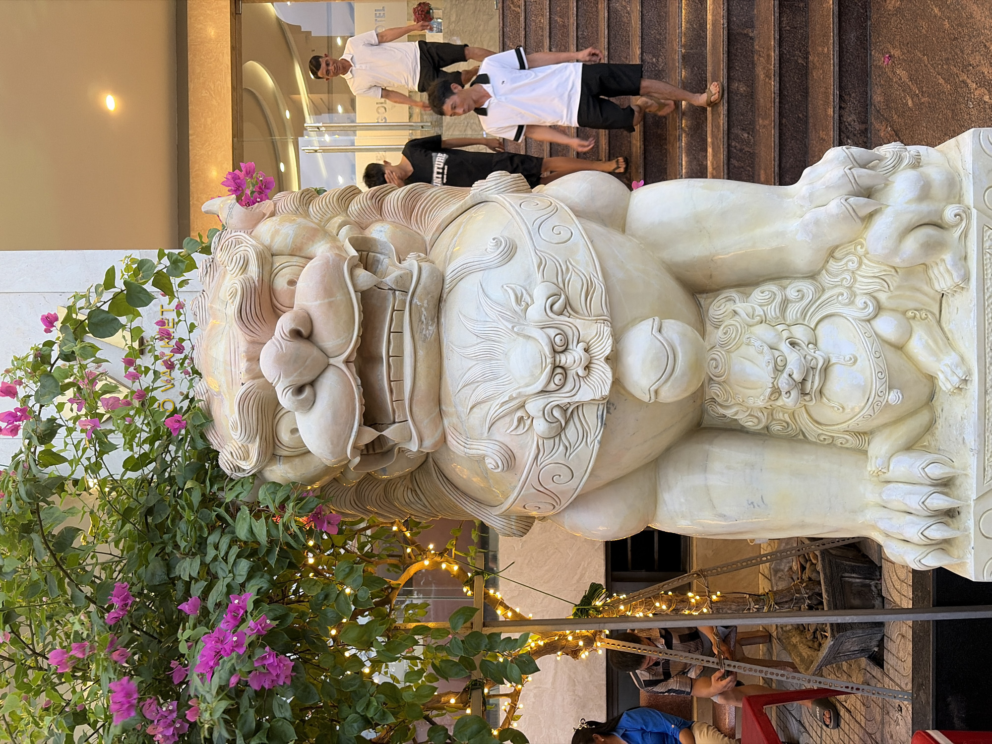 An ornate stone sculpture of a Foo dog, a mythical Chinese guardian lion, stands prominently in front of a building entrance, partially obscured by a tree adorned with pink bougainvillea flowers and string lights. The sculpture is detailed, featuring intricate carvings of a lion's head, a decorative collar, and a smaller lion at its base. To the right of the sculpture, a set of stone stairs leads up to an arched entryway with glass doors. Three young men are descending the stairs, two wearing white polo shirts and black shorts, and one in a black t-shirt and shorts. A fourth man, in a white polo shirt and black shorts, stands at the top of the stairs near the entrance, holding a bouquet of pink flowers. On the left side of the image, two women are seated in chairs next to a table, with a potted plant near the Foo dog sculpture. The overall setting appears to be the entrance to a hotel or establishment in Đà Nẵng, Vietnam.