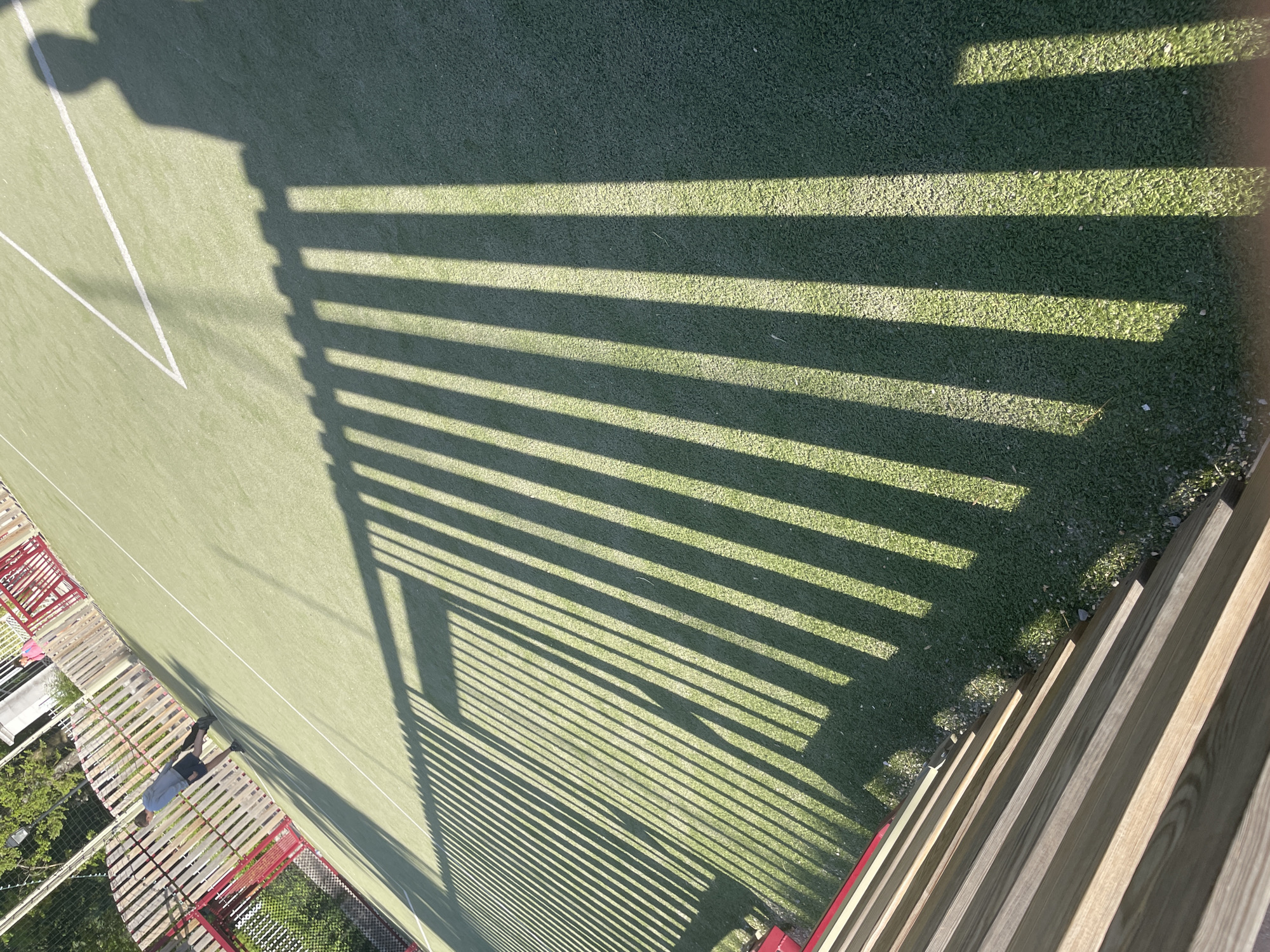 An outdoor artificial turf soccer field is pictured from a high angle, casting long, striped shadows from a wooden fence. In the background, a person in a blue hoodie and black shorts walks away from the camera on the field. The fence in the foreground is made of light-colored wooden planks.