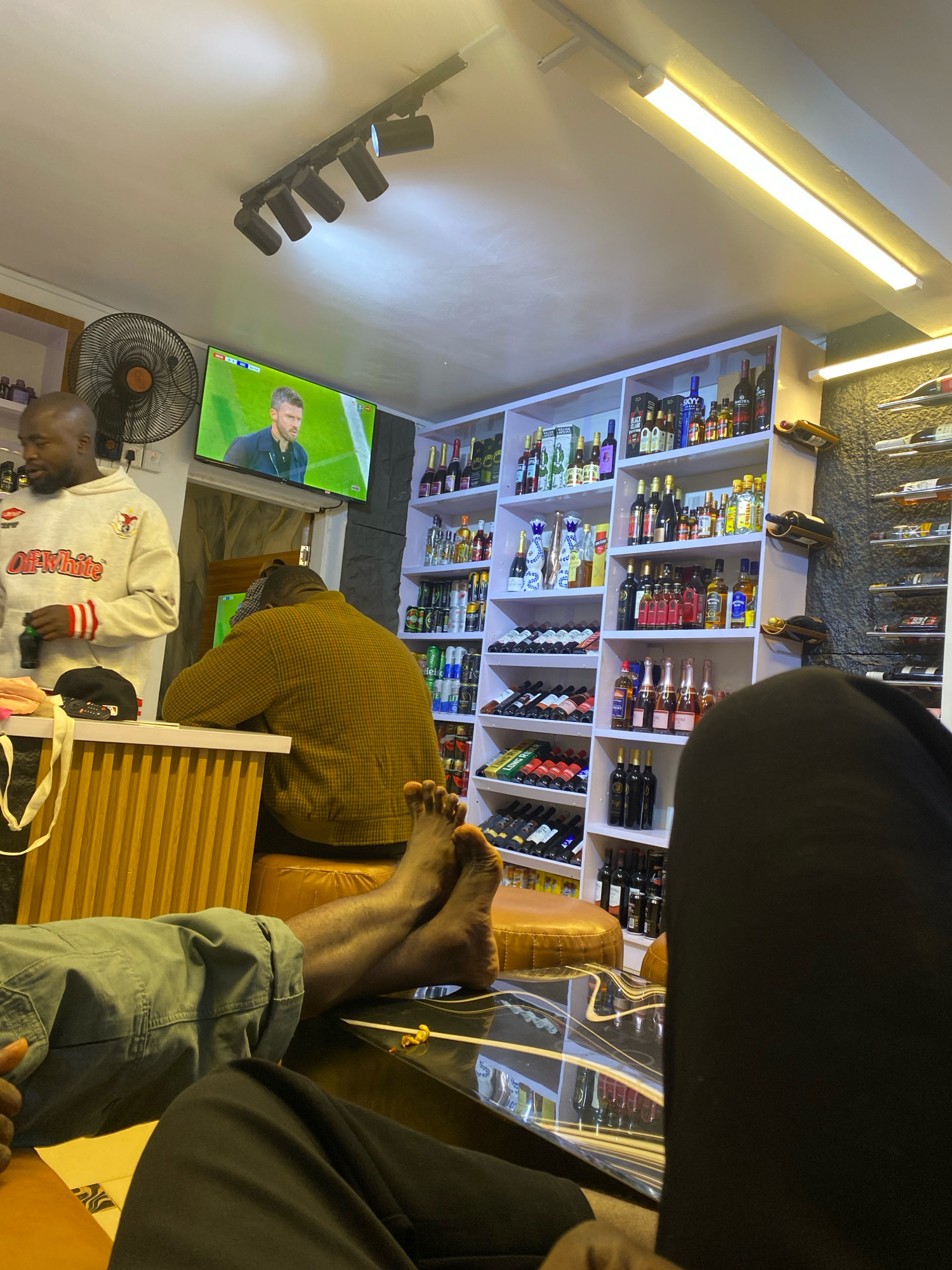 The image depicts the interior of a bar or lounge in Jos, Nigeria. In the background, shelves are stocked with various bottles of alcohol, suggesting a retail or service establishment. A television screen mounted on the wall displays a soccer match, with a player visible on screen. A man in a light-colored hoodie with red and white stripes stands near the television, holding a bottle. Another man, facing away from the camera, is seated at a counter. In the foreground, two individuals are visible, with one person's legs and feet extended towards the camera, resting on a reflective surface that appears to be a table or counter. The lighting in the establishment is a mix of overhead track lighting and a long fluorescent tube light. The overall atmosphere suggests a casual gathering where patrons are watching sports and possibly consuming beverages.