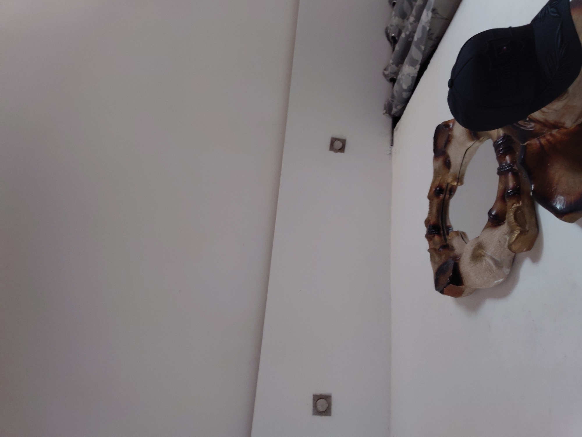 The image displays an indoor scene featuring a large, abstract wooden sculpture mounted on a white wall. The sculpture exhibits rich, polished wood tones ranging from dark brown to lighter beige, with an intricate, organic, and somewhat gnarled form. A black baseball cap rests on a prominent part of the sculpture, suggesting it might be stylized as a head or bust.
To the right of the sculpture, partially visible in the upper right corner, is a curtain or fabric with a distinctive grey and black camouflage pattern. The white wall, which serves as the primary background, features two small, square, metallic-rimmed objects with circular centers, possibly recessed lights or electrical outlets, embedded into its surface. One is positioned roughly midway up the wall, and another lower down.
The scene appears to be static, showcasing a decorative arrangement in a room. There are no people visible, and no specific activity is taking place. The lighting is soft and even, indicating an indoor setting. No discernible text is present in the image. While the provided context suggests Santa Ana, Venezuela, no specific visual cues within the image confirm this location.