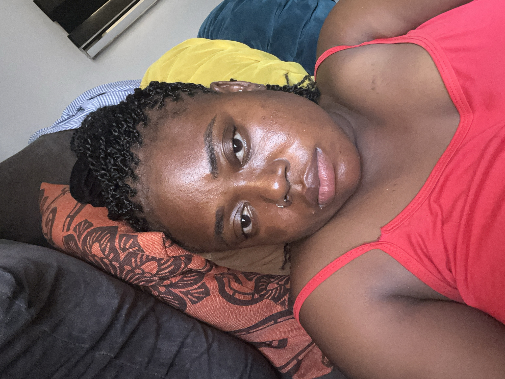 A close-up, waist-up shot of a young Black woman lying on a bed. She is wearing a red tank top. Her hair is styled in small braids. Her face is turned slightly towards the camera. She has a nose ring. She is lying on a pillow with a black and orange patterned cover. In the background, there are pillows of blue and yellow fabric. A black and silver object, possibly a projector, is mounted on the wall in the upper left corner. The lighting suggests it is daytime. The overall mood is relaxed and casual. No visible text is present in the image.