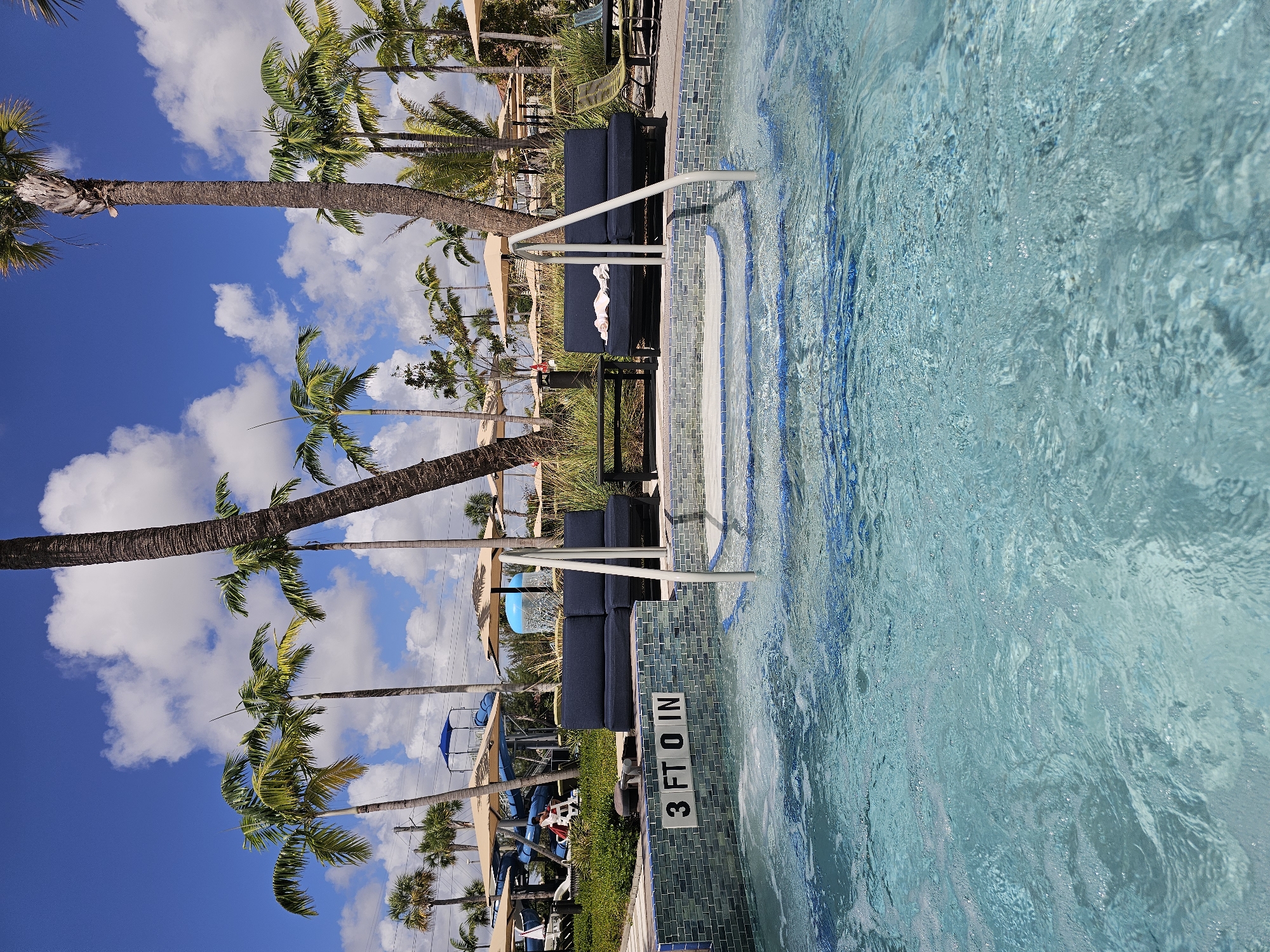 The image depicts a swimming pool, likely at a resort or hotel in Miami, United States of America, indicated by the presence of palm trees and a clear blue sky with puffy white clouds. The pool's depth is marked as "3 FT 0 IN" on the tiled wall. A set of white metal pool stairs provides access to the water. Lounge furniture is visible beside the pool, featuring dark cushions and additional tables. In the background, a water slide structure can be seen, further suggesting a resort or recreational setting. No people are prominently featured, but the presence of chairs suggests it is an area intended for guests.