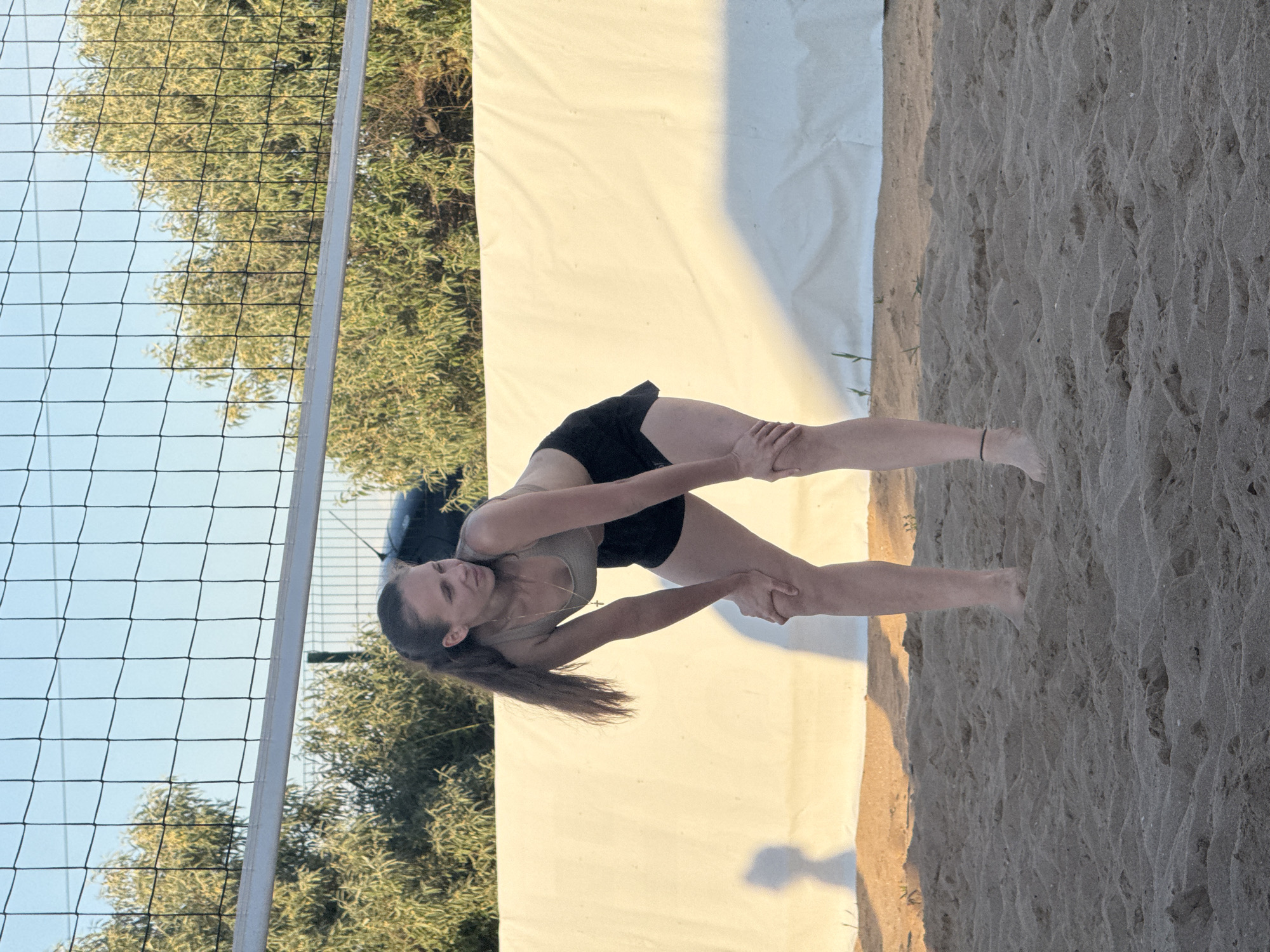A woman is standing on a sandy beach, bent at the knees with her hands on her thighs. She is wearing a grey sports bra and black shorts, and a necklace with a cross pendant. A volleyball net is visible behind her, with trees and a light-colored fabric backdrop beyond that. The sky is clear and blue. The location is identified as Shchelkino, Ukraine.