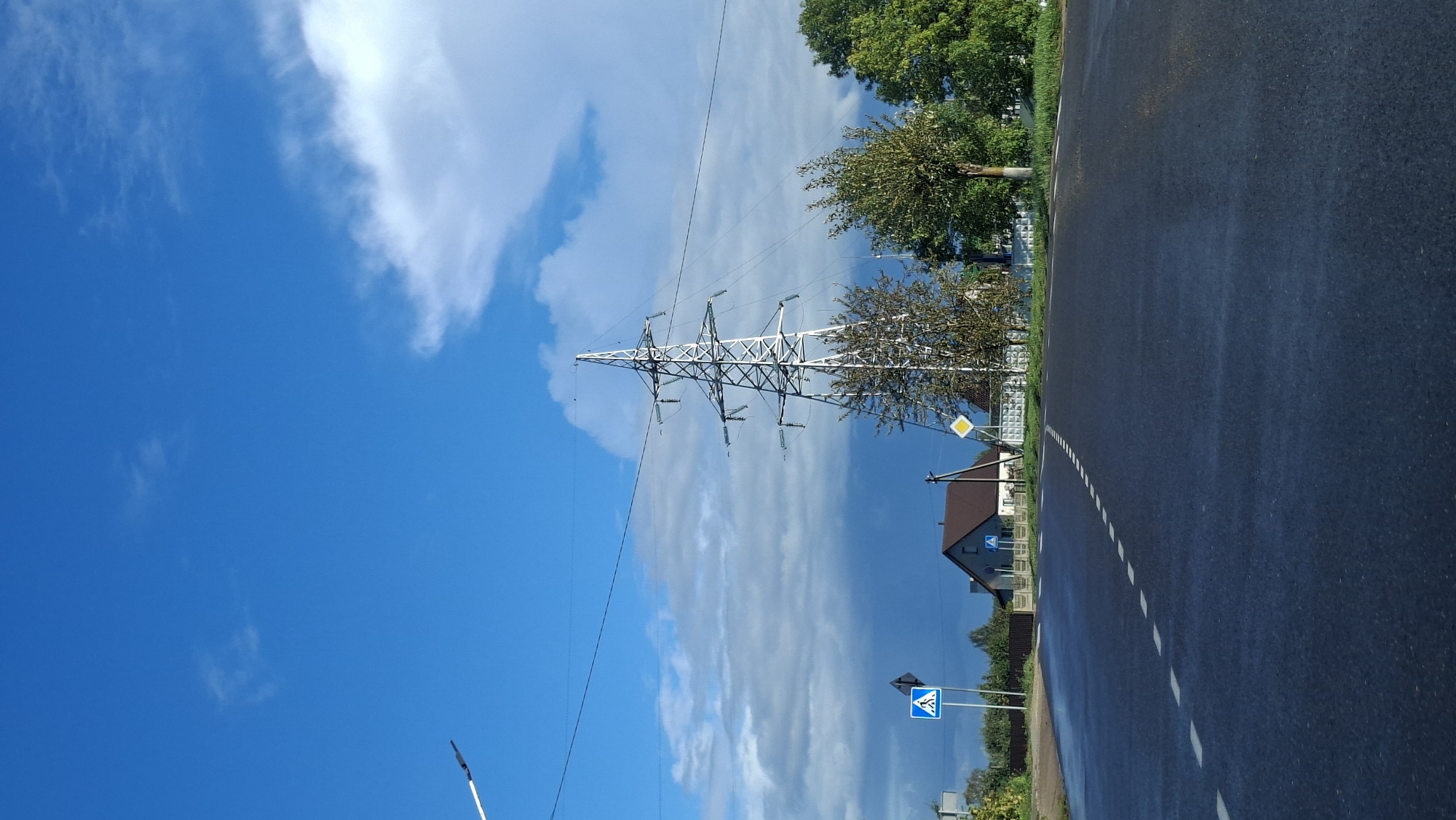 The image depicts a rural road in Myadzyel, Belarus, on a partly cloudy day. A paved road with a dashed white line runs through the foreground, appearing slightly wet. On the left side of the road, there is a pedestrian crossing sign and a fence. Beyond the fence, a building with a brown roof is visible, as well as trees. Dominating the mid-ground is a tall, silver electricity pylon with multiple crossbars and wires extending from it. To the right of the pylon are several trees with green and yellow foliage, suggesting it may be autumn. The sky is a vibrant blue with scattered white and dark clouds.
