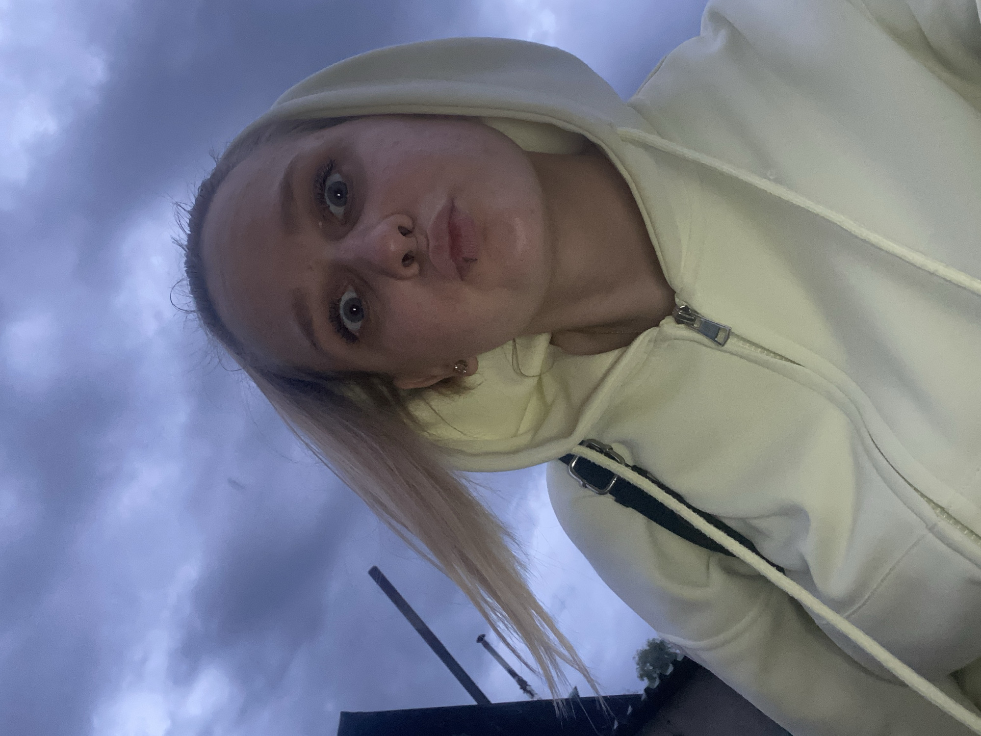A woman with blonde hair pulled back into a ponytail is taking a selfie outdoors under a cloudy sky. She is wearing a white hooded sweatshirt with a zipper. Her lips are pursed in a slight pout. In the background, there are glimpses of street elements, suggesting an urban setting. The specific location is the city of Chaikovsky, Russia.