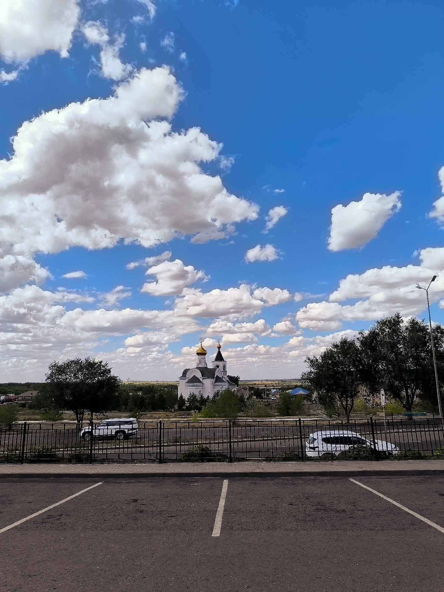 The image displays a wide-angle view of a landscape featuring a white church with golden domes prominently in the center background. The sky is a vibrant blue, filled with numerous fluffy white clouds. In the foreground, there is a paved parking lot with white parking lines. A black metal fence runs horizontally across the mid-ground, separating the parking lot from a grassy area with scattered trees and shrubs. Beyond the fence, two white vehicles are visible, one a sport utility vehicle and the other a sedan. The church is situated amidst a sparse treeline and buildings, suggesting it is located within a town or city. The overall scene suggests a sunny day with a clear sky and a few scattered clouds. The architecture of the church and the surrounding landscape are consistent with a location in Kazakhstan, potentially near the city administration of Qarağandı.