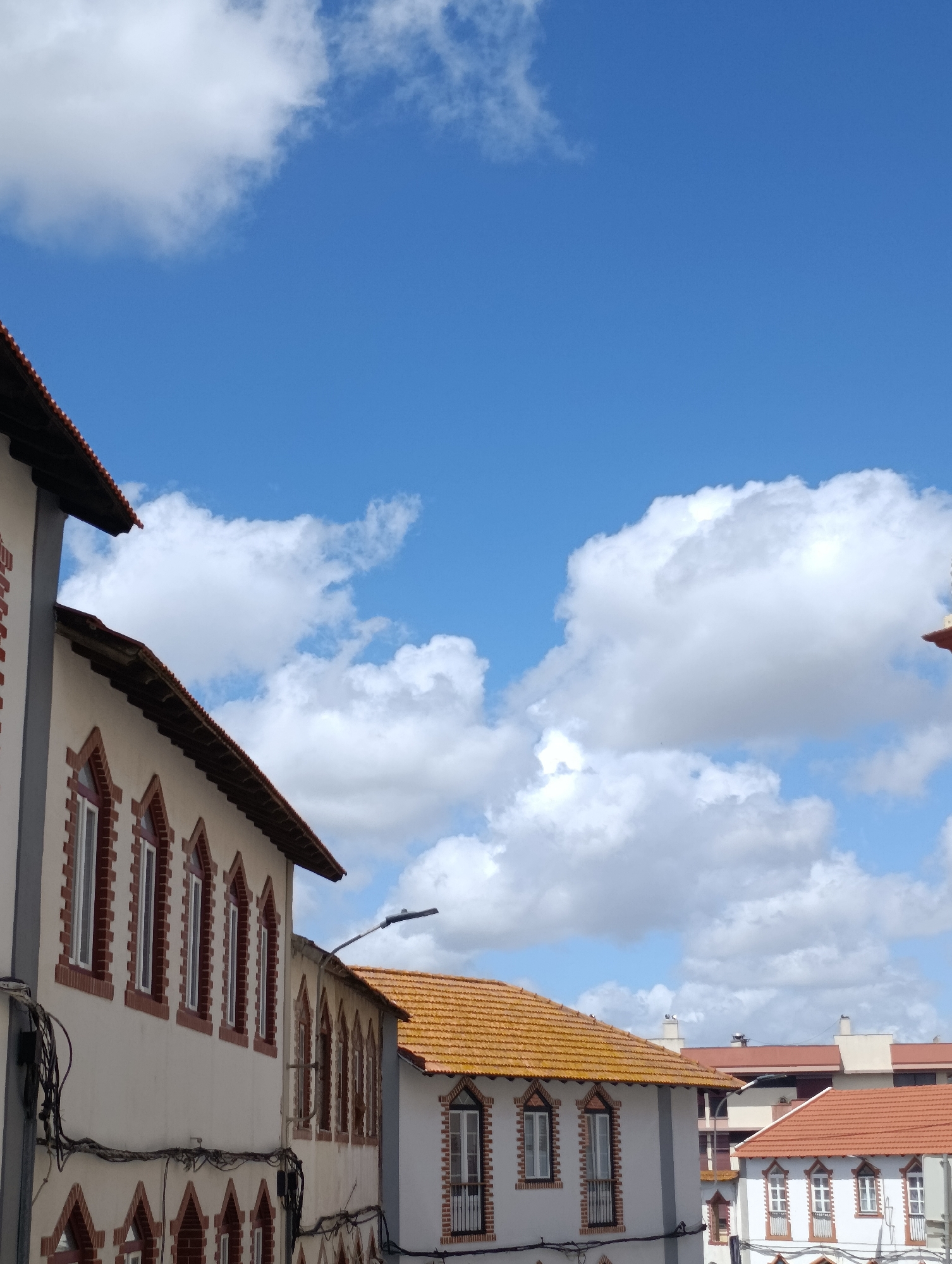 This is an outdoor, eye-level, vertical photograph taken from a low angle. It shows the upper facades of several buildings with traditional Portuguese architecture, likely in Setúbal, Portugal. The buildings feature light-colored walls, dark-tiled roofs with visible orange tiles, and distinctive arched windows framed by red brick. The windows are multiple and evenly spaced along the building's walls. Some electrical wires are visible running along the building facades. In the background, there are more buildings with similar architectural elements and red roofs. The sky is a clear, bright blue with scattered, puffy white clouds. A street lamp is visible on a pole. The image captures a sunny day with good visibility. There are no people or vehicles visible in the frame.