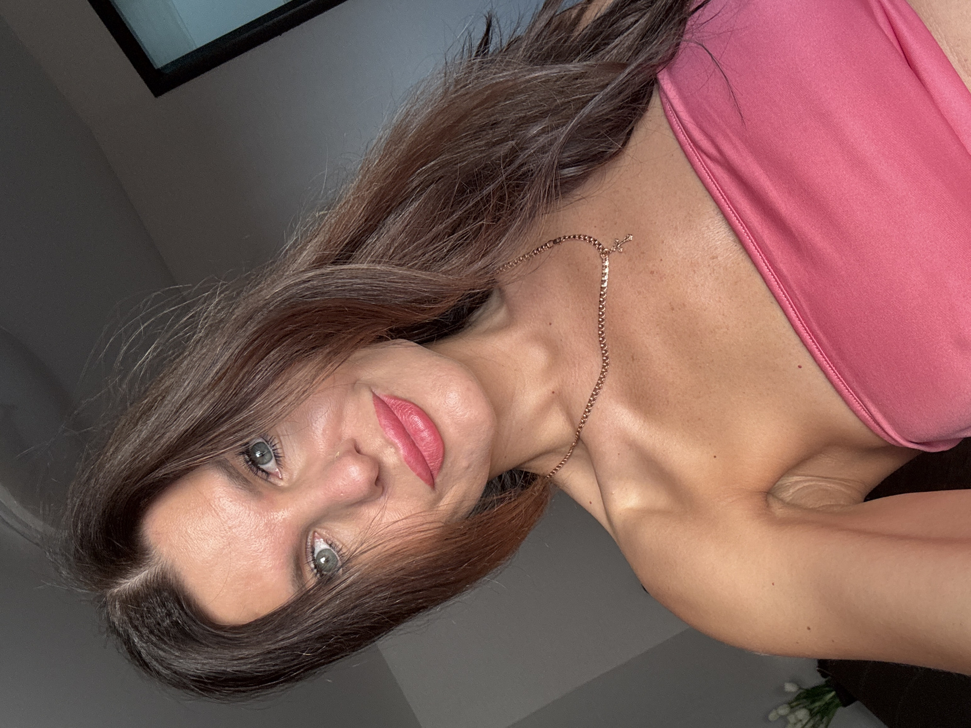 The image shows a close-up selfie of a woman with long, dark brown hair and blue eyes. She is wearing a pink strapless top and a delicate gold necklace with a cross pendant. Her lips are painted a bright pink. The background is a muted gray wall with a shadow from a window. There are no discernible elements indicating the location in Chaikovsky, Russia.