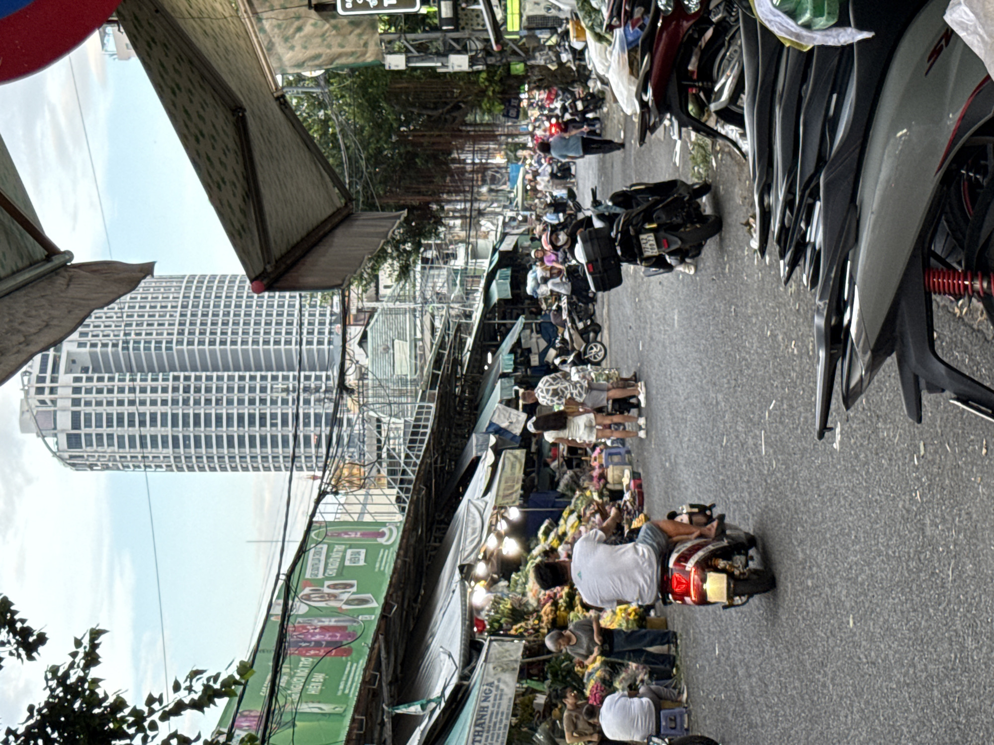 The image captures a busy street scene in Nha Trang, Vietnam, likely a marketplace, with a tall, modern skyscraper dominating the background. The foreground is filled with a variety of motorcycles parked and in motion, typical of Southeast Asian cityscapes. Several stalls line the street, displaying a multitude of flowers, suggesting a flower market. People are seen browsing, shopping, and riding their motorbikes. The atmosphere appears lively and bustling, with awnings providing shade over the market stalls. The sky is overcast.