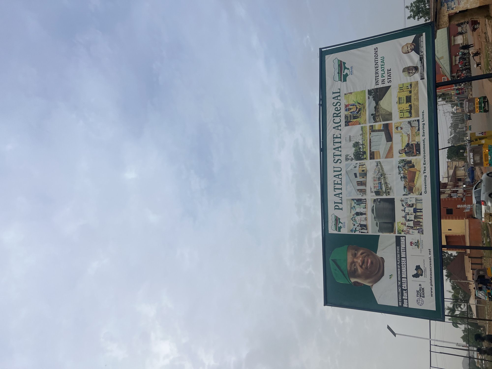 The image depicts a billboard in Bukuru, Nigeria, advertising Plateau State ACRESAL and the interventions in Plateau State. The billboard features images of Governor Caleb Manasseh Mutfwang and Mr. Garba Gowon Gonkol. The background shows a street scene with buildings, vehicles (including yellow three-wheelers), and pedestrians.