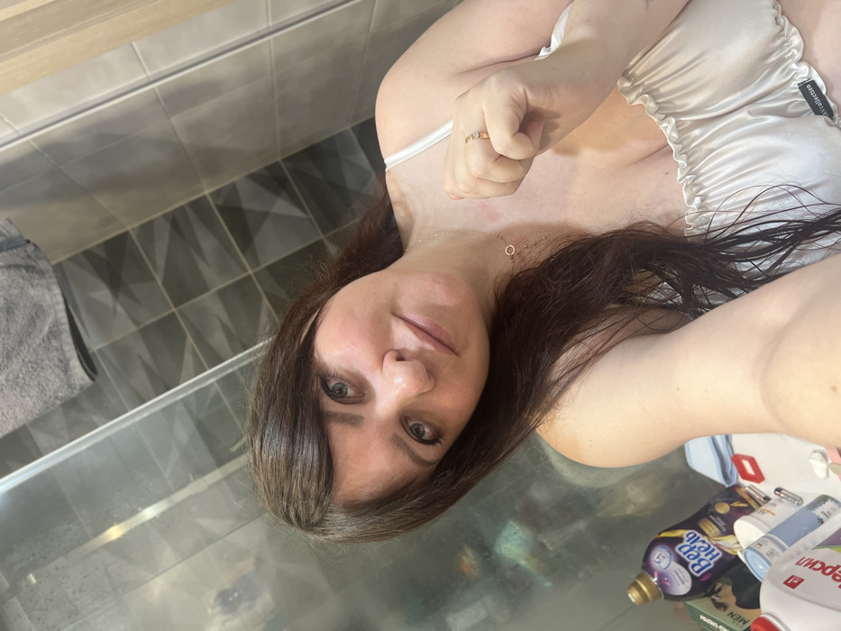 A female individual is depicted in a selfie within a bathroom in the city of Noviy, Russia. She has long dark brown hair, light eyes, and wears a light-colored, ruffled satin top with an "AV Collection" tag. A thin gold necklace with a circular pendant is visible on her neck, where a faint reddish mark is present. Her right hand, raised in a loose fist, displays a gold ring with a clear stone on her ring finger. The background features a dark grey tiled wall with a geometric pattern, partially obscured by a reflective glass surface, likely a shower door. A grey towel hangs in the upper right. On a counter to the lower left, a purple bottle of "ВерНель" (Vernel) fabric softener and a white bottle of "Персіл" (Persil) laundry detergent are visible, along with other miscellaneous items.