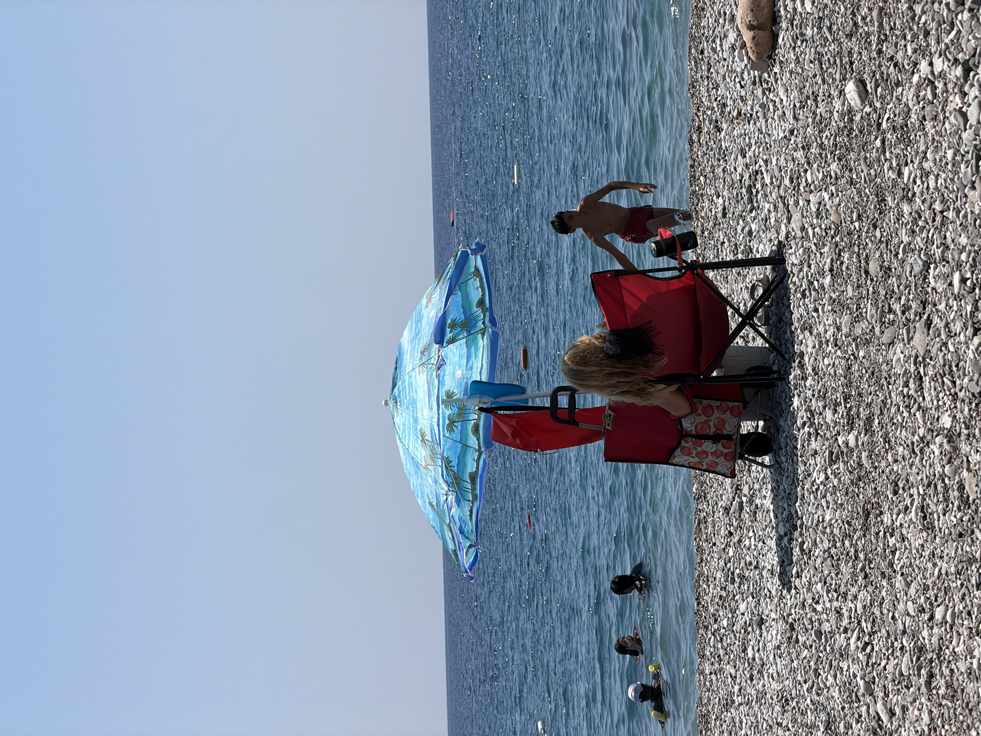 The image shows a sunny day at a pebbly beach in Kemer, Turkey. In the foreground, two red beach chairs are set up under a blue umbrella decorated with palm trees. One chair is occupied by a person from behind, facing the sea, with their blonde hair visible. Next to this chair is a red rolling cart with a floral pattern. To the right of the umbrella, a young boy in red swim trunks is walking out of the sea. In the middle ground, several people are swimming in the clear blue water. The sea stretches to the horizon under a clear blue sky. The beach is covered with smooth, grey and white pebbles. A large, light-colored rock is visible on the right side of the beach.